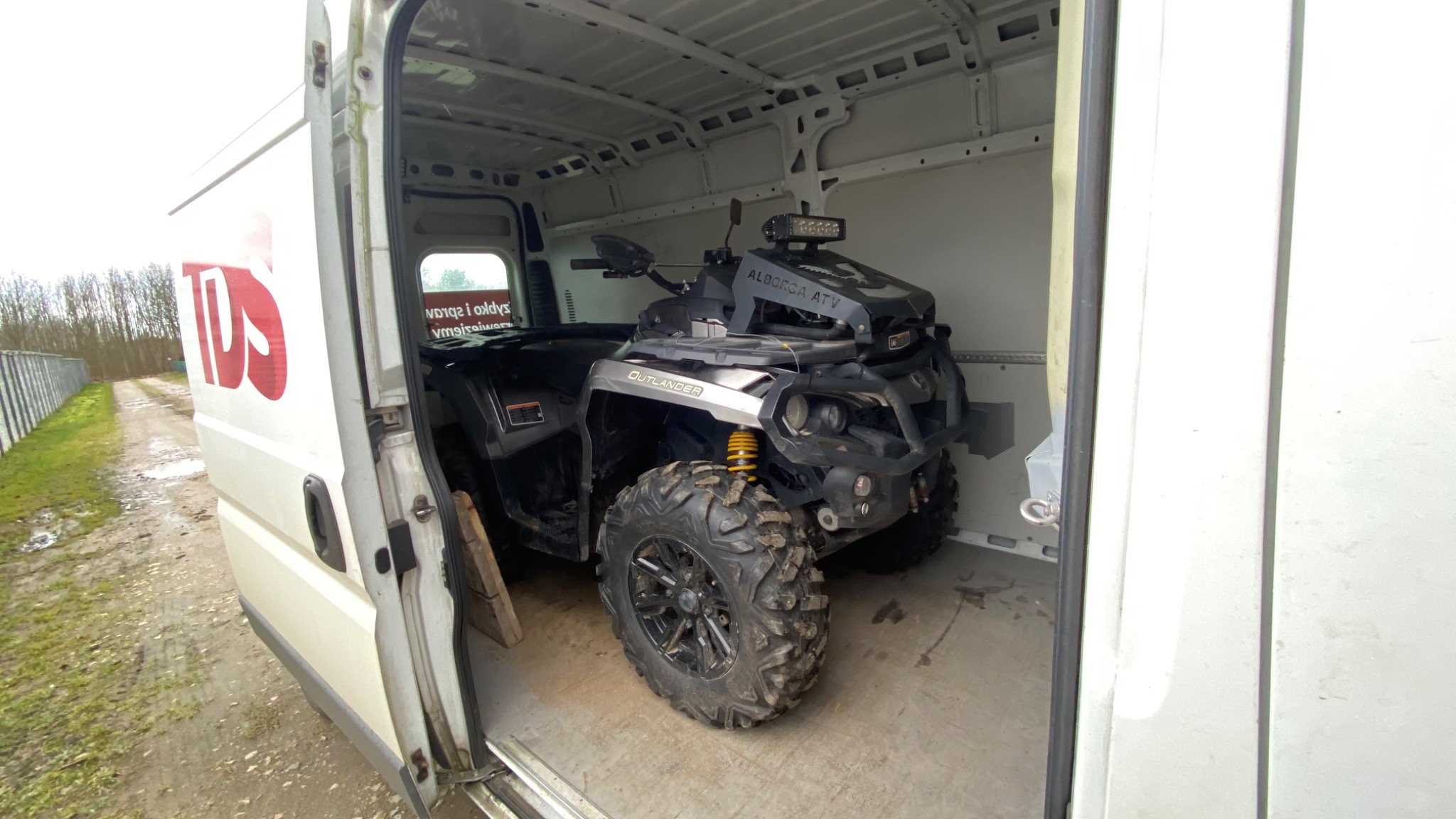 Czarny quad ATV marki Alborca w przestrzeni ładunkowej białego busa, widoczne duże opony terenowe i żółte amortyzatory.