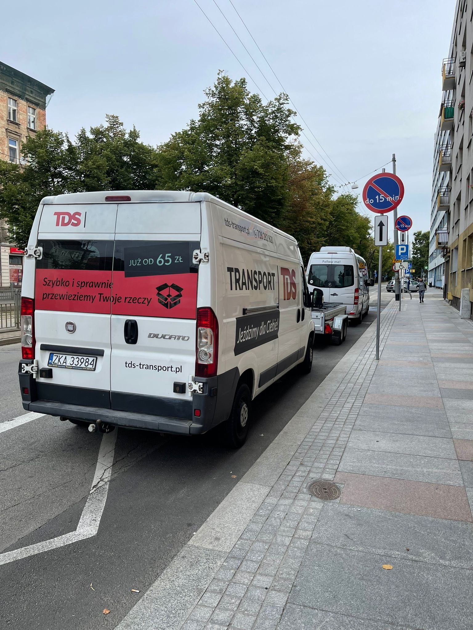 Biały bus firmy transportowej TDS z przyczepką zaparkowany na ulicy miejskiej. Na tylnych drzwiach reklama: 'Szybko i sprawnie przewieziemy Twoje rzeczy, już od 65 zł'.