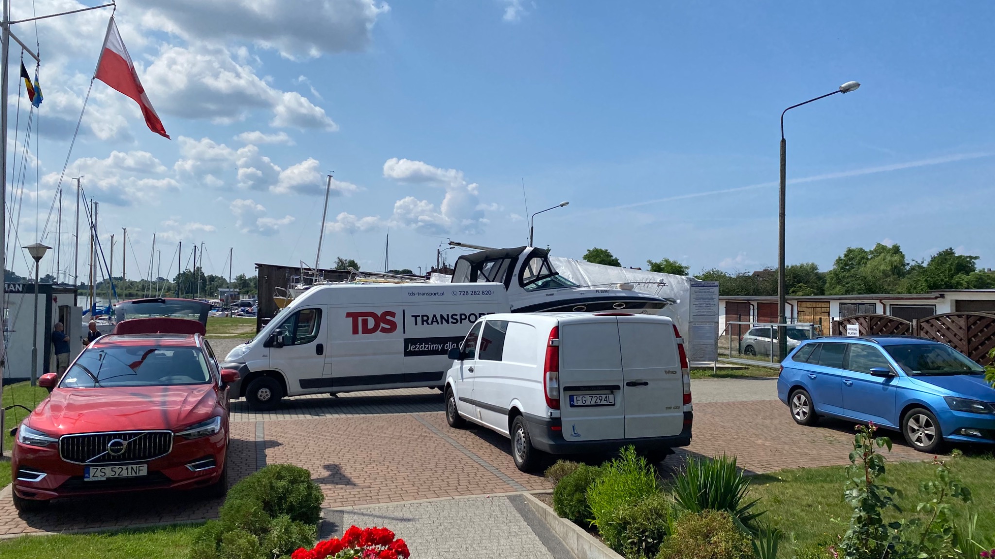 Dwa busy transportowe, biały Mercedes Vito i biały Fiat Ducato z logo firmy, stoją na tle mariny z łodziami, pod polską flagą.