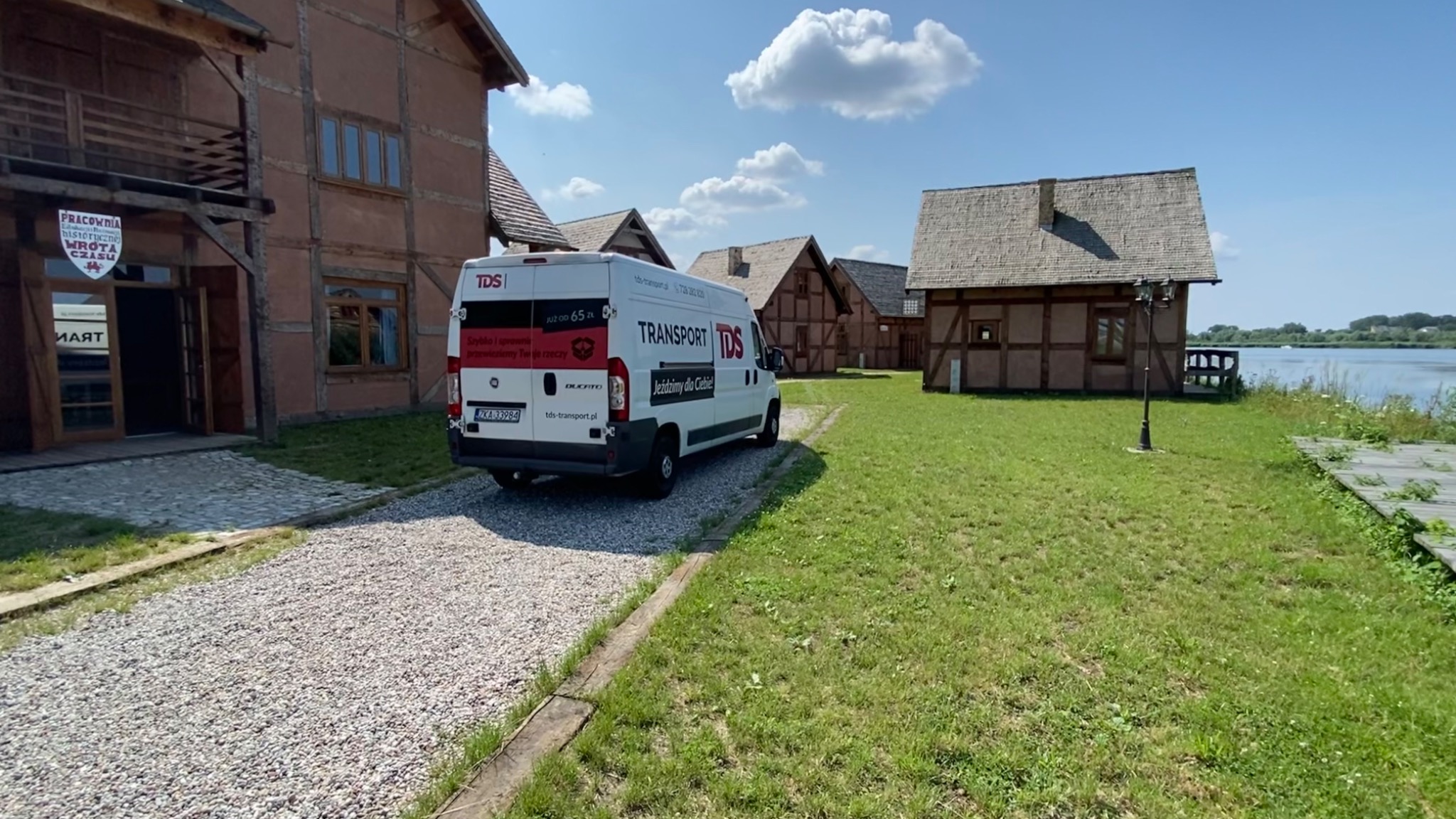 Biały bus transportowy z logo firmy TDS zaparkowany na żwirowej drodze przed budynkami w stylu skansenowym, w tle widoczne jezioro.