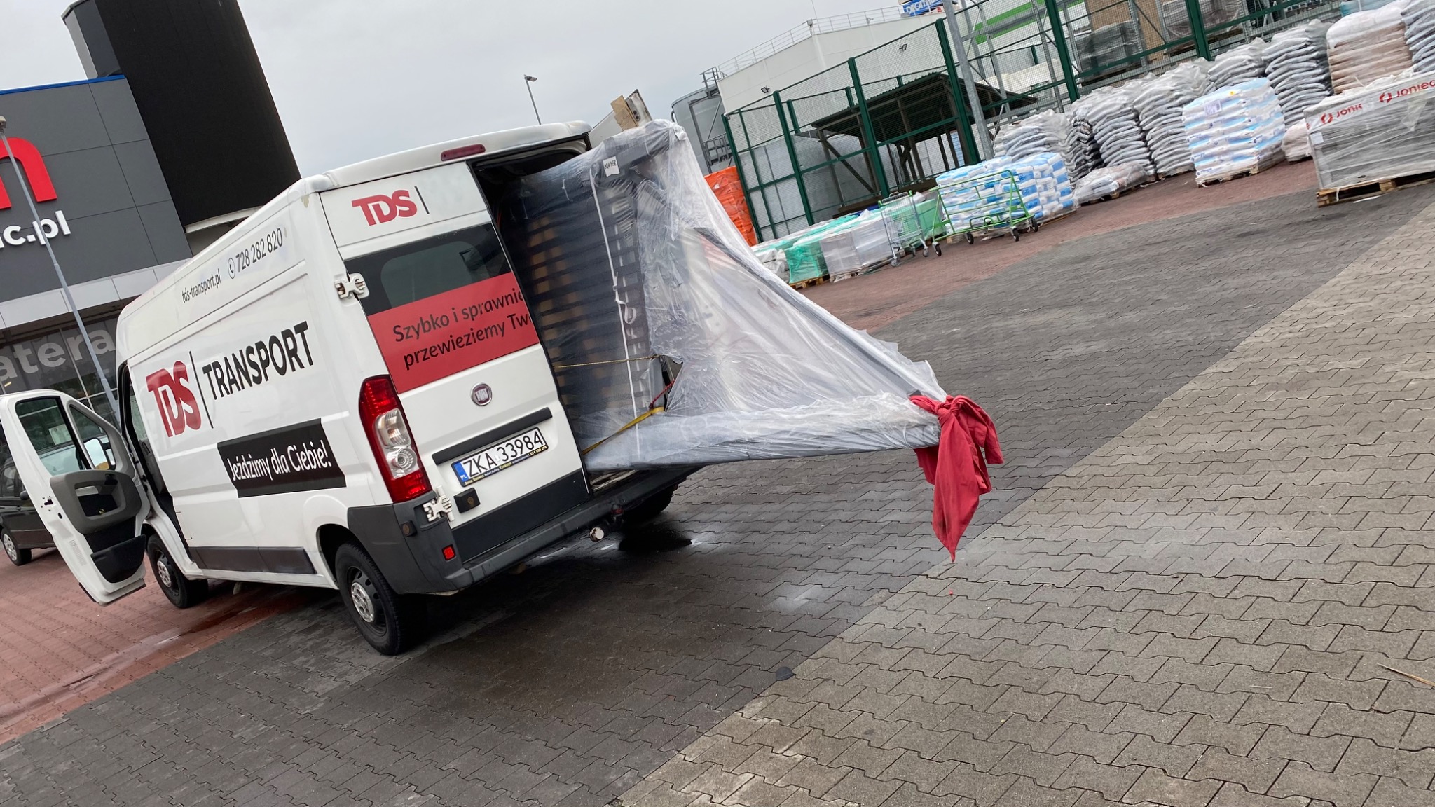Biały bus firmy transportowej TDS z otwartymi tylnymi drzwiami, przewożący długi, owinięty folią i zabezpieczony czerwoną szmatą ładunek, zaparkowany na parkingu sklepu budowlanego.