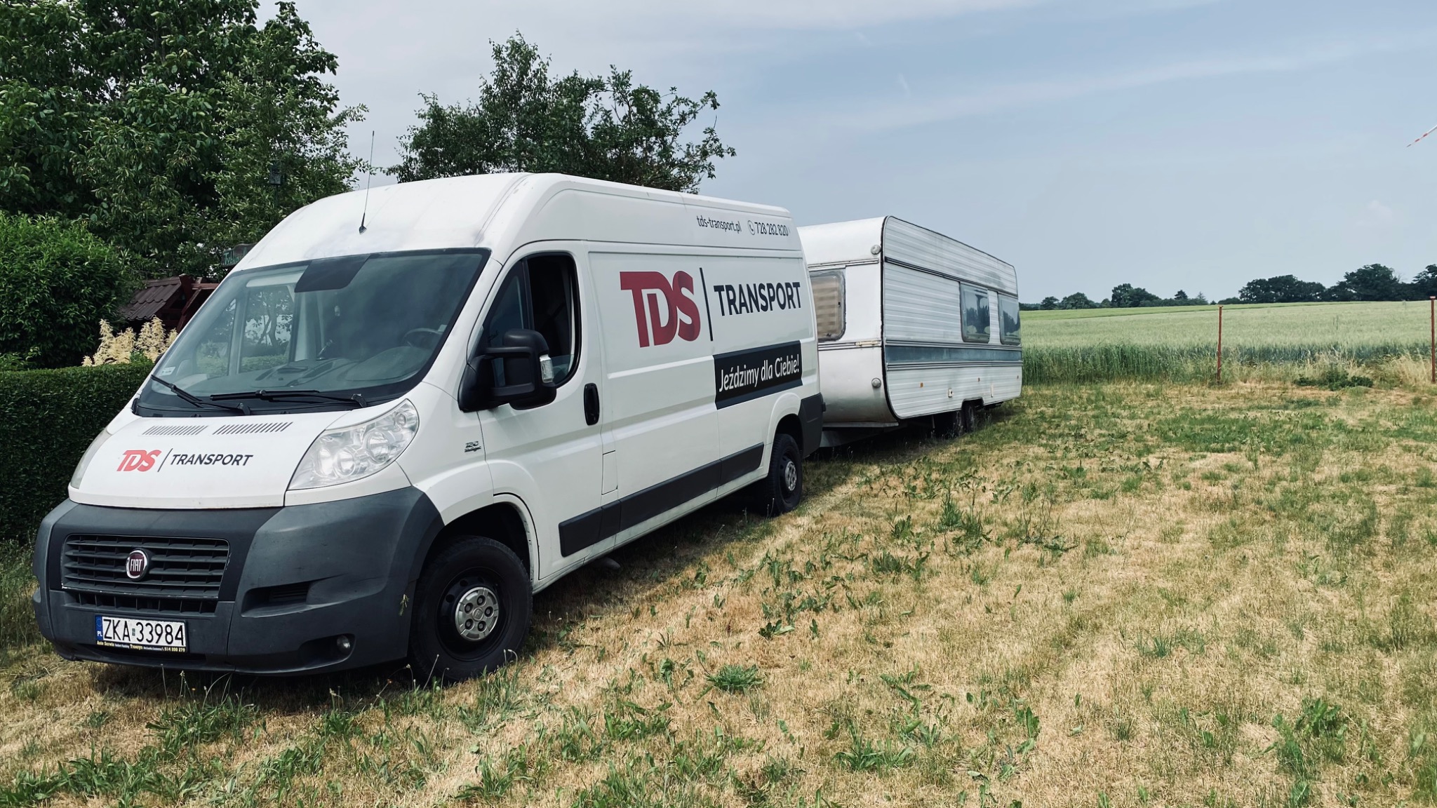 Biały bus z logo TDS Transport ciągnie przyczepę kempingową na tle pola i drzew. Na busie widoczny numer telefonu.