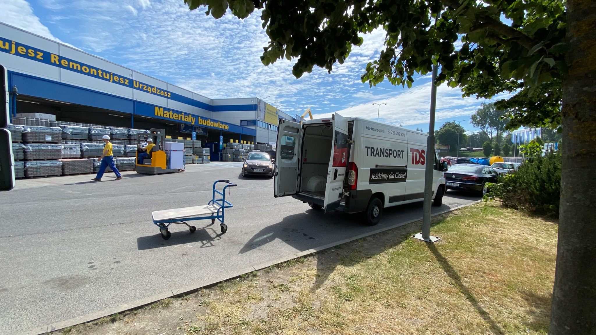 Biały bus z otwartymi drzwiami, z napisem TRANSPORT TDS, zaparkowany przed marketem budowlanym w Wolinie, z wózkiem transportowym na pierwszym planie i pracownikami marketu obsługującymi wózek...