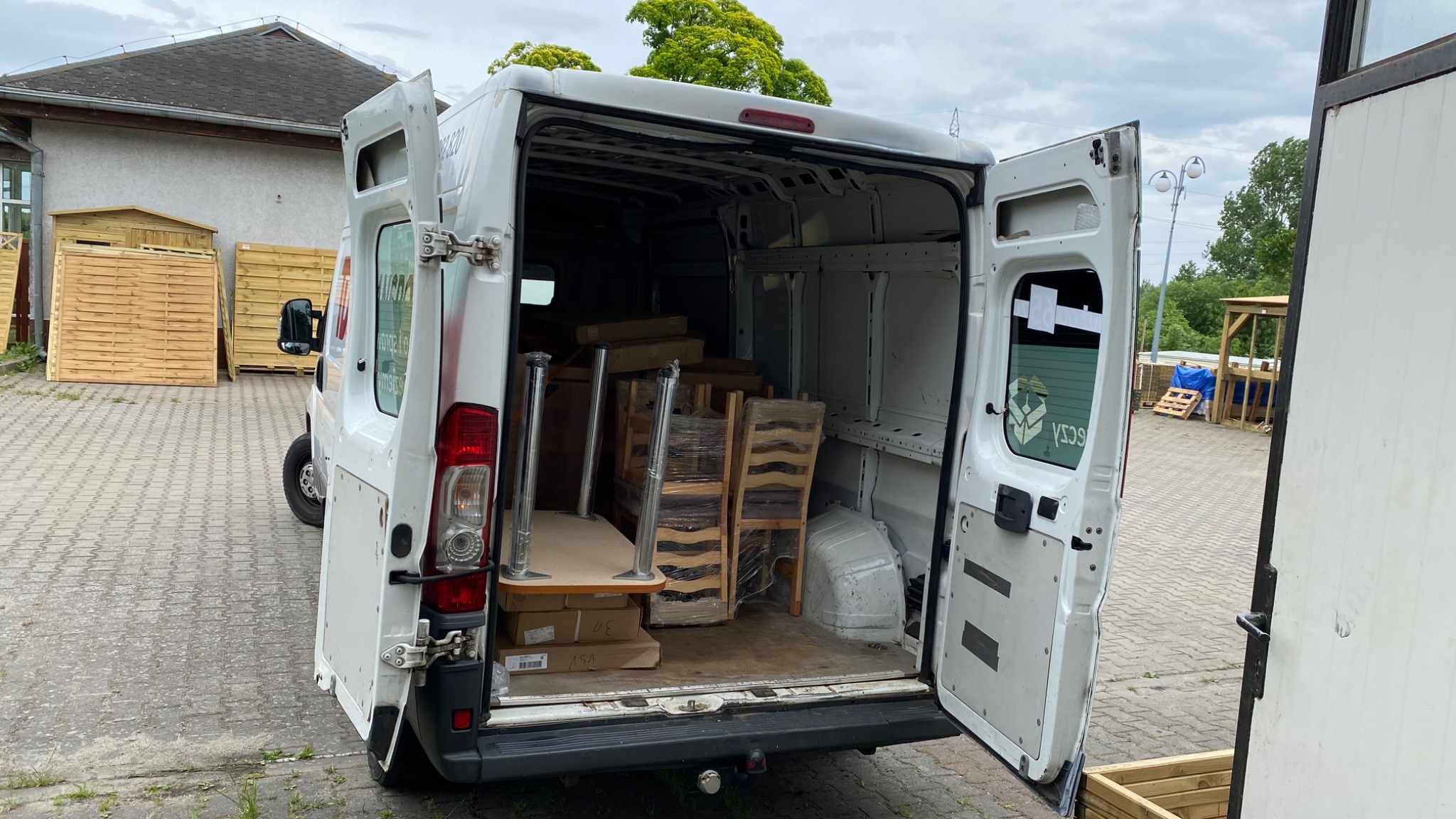 Otwarty tył białego furgonetki załadowany stołami i krzesłami owiniętymi folią stretch, na zewnątrz widoczne drewniane panele ogrodzeniowe i kostka brukowa.