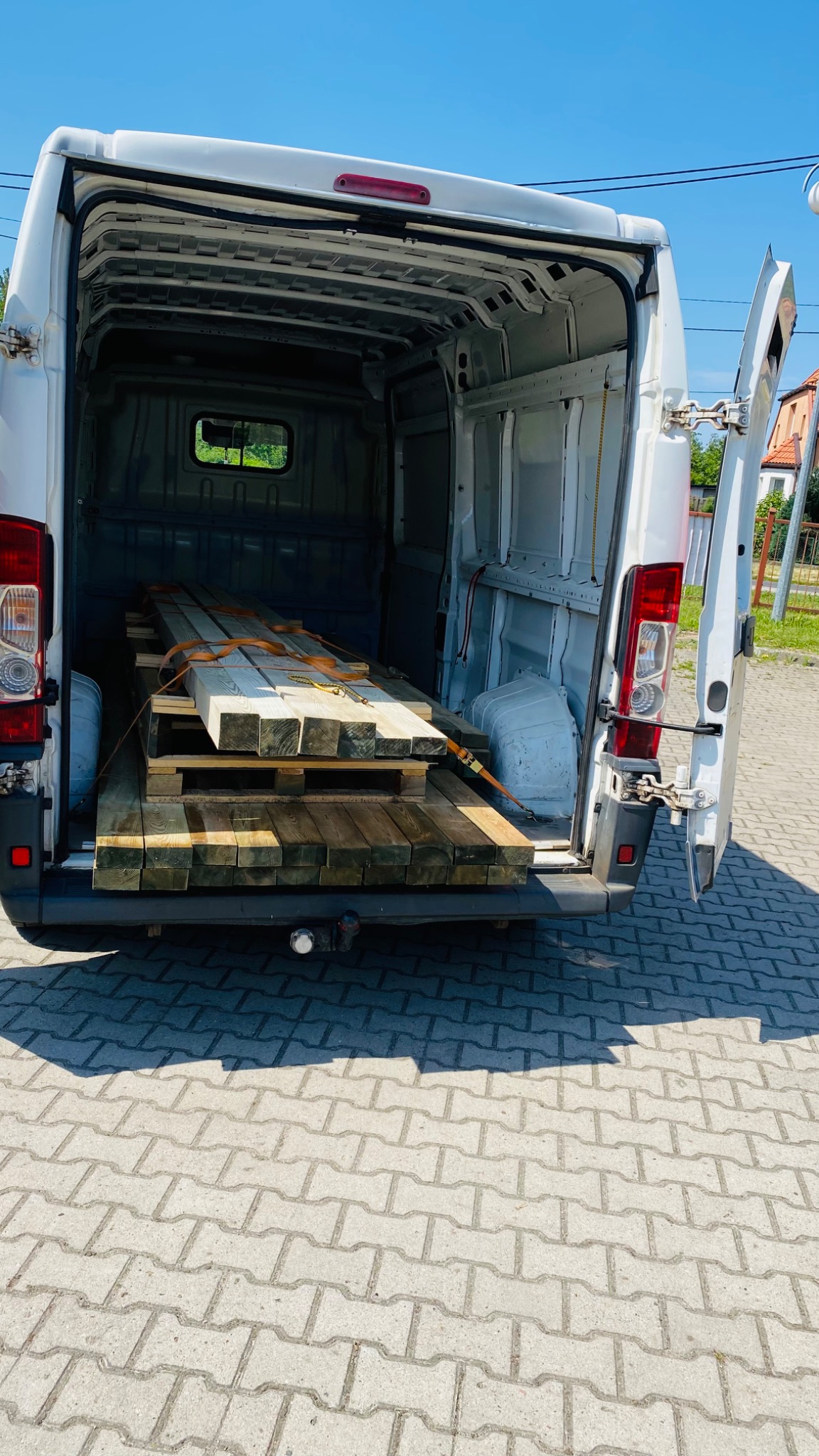 Wnętrze białego busa z otwartymi tylnymi drzwiami, załadowane paletą z drewnianymi belkami zabezpieczonymi pomarańczowymi pasami.