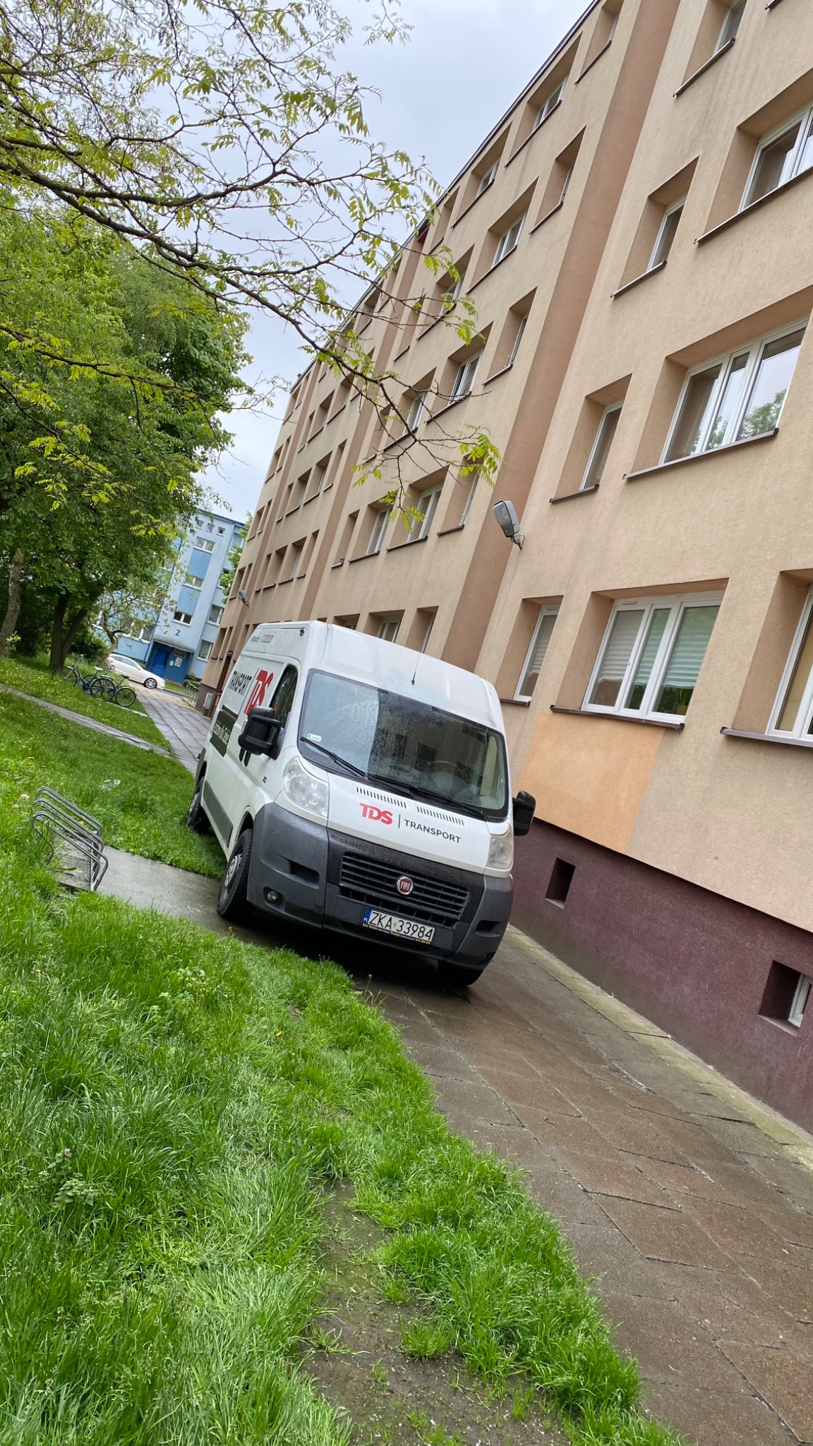 Biały bus firmy transportowej TDS Transport zaparkowany na chodniku przed blokiem mieszkalnym w pochmurny dzień.