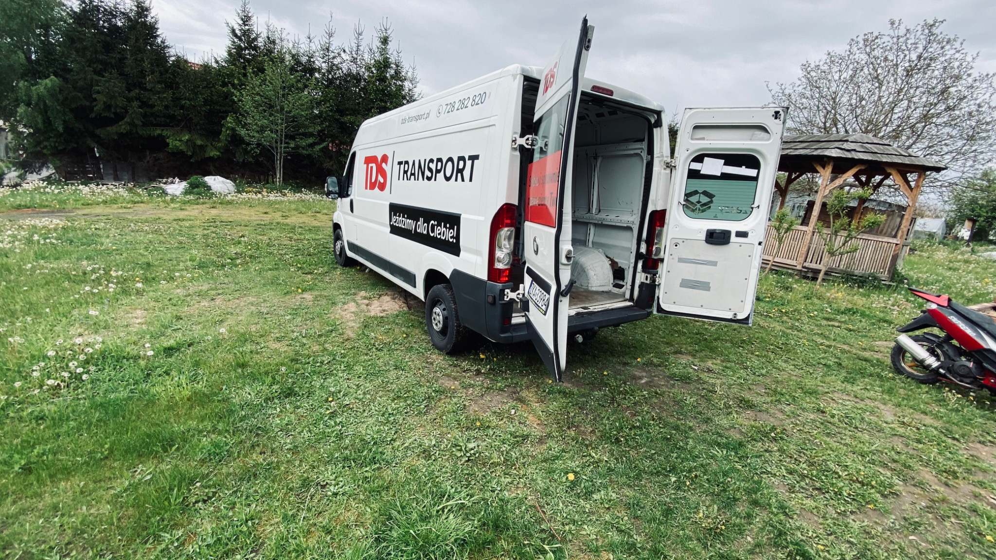 Biały samochód dostawczy z otwartymi drzwiami bagażnika, zaparkowany na trawiastym terenie z widoczną altanką w tle i skuterem po prawej stronie.
