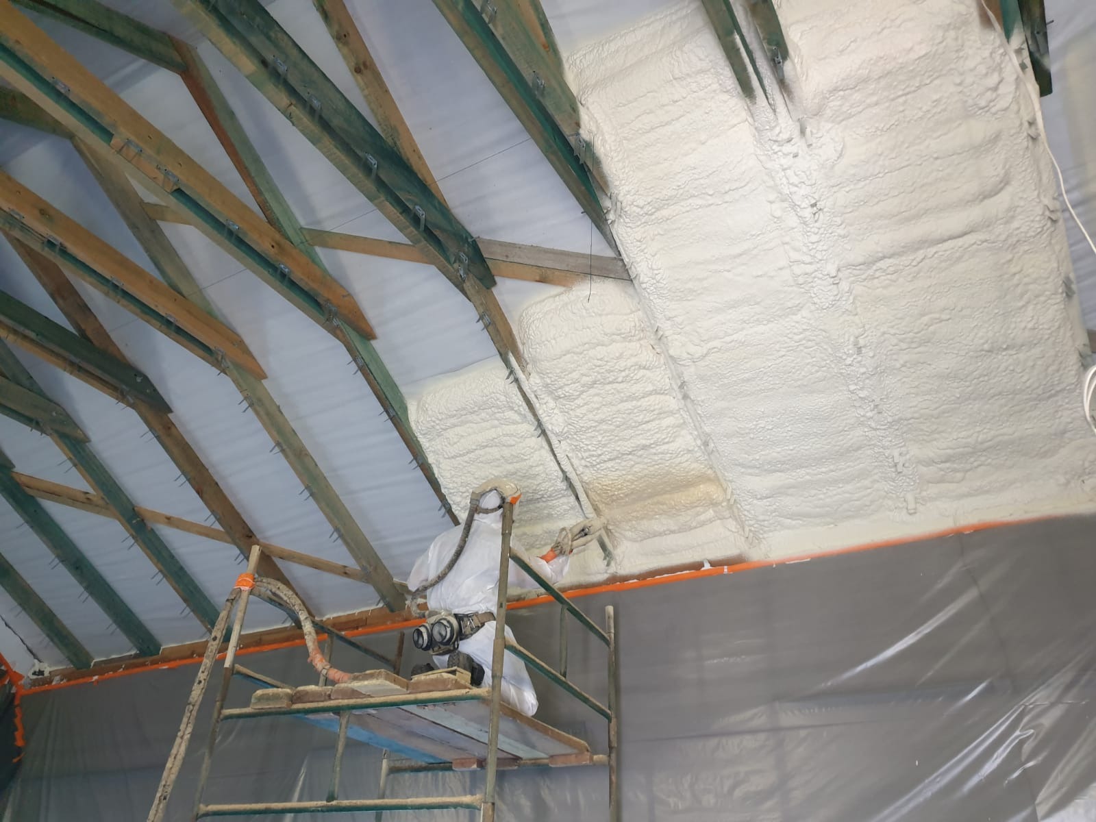 Pracownik w kombinezonie ochronnym aplikuje piankę izolacyjną na poddasze z widoczną drewnianą konstrukcją dachu, stojąc na rusztowaniu.