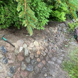 Kacper Czaja - Skarpa obłożona kamieniami polnymi, widoczny fragment zieleni i narzędzie ogrodnicze.