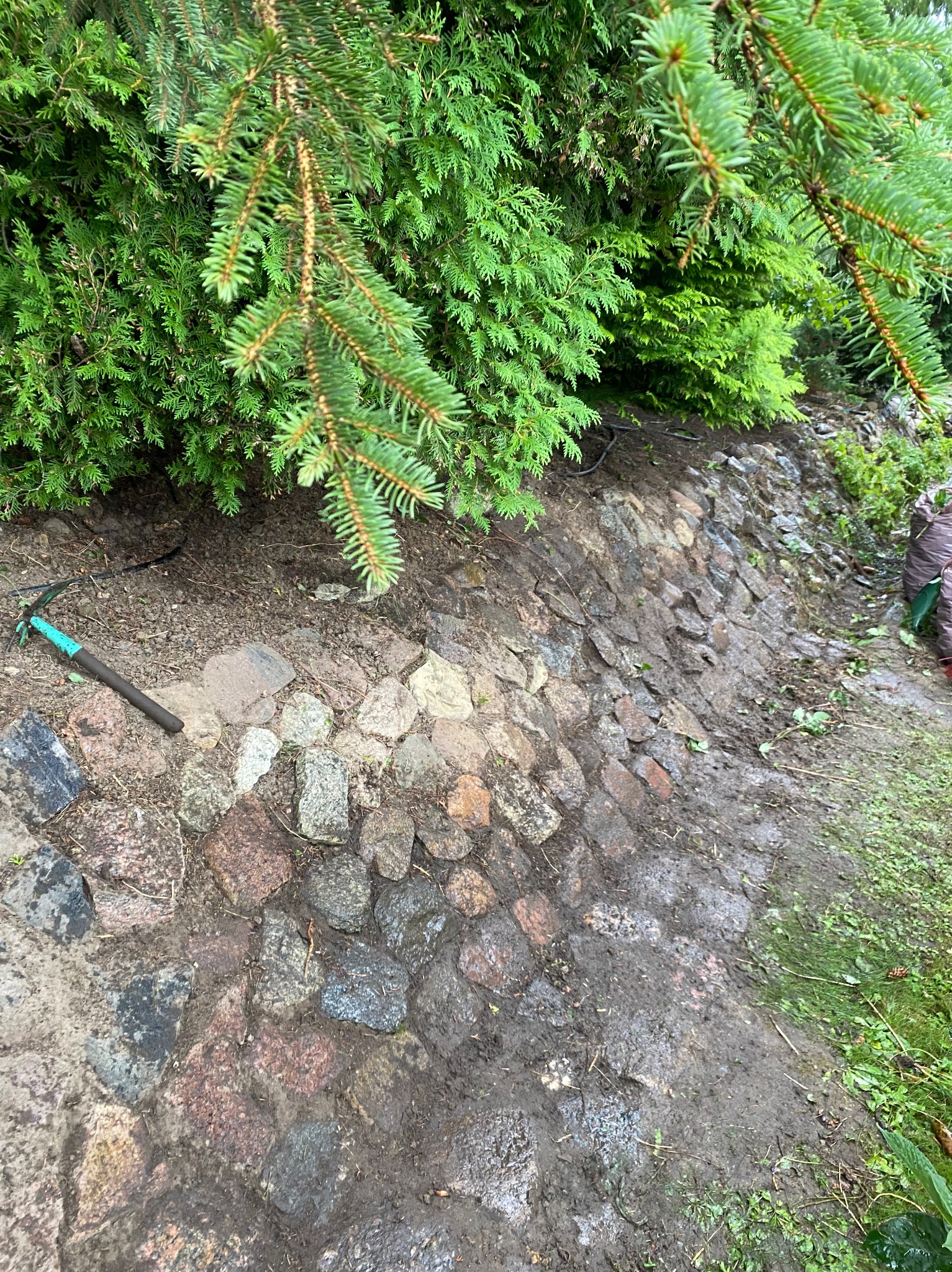 Skarpa obłożona kamieniami polnymi, widoczny fragment zieleni i narzędzie ogrodnicze.