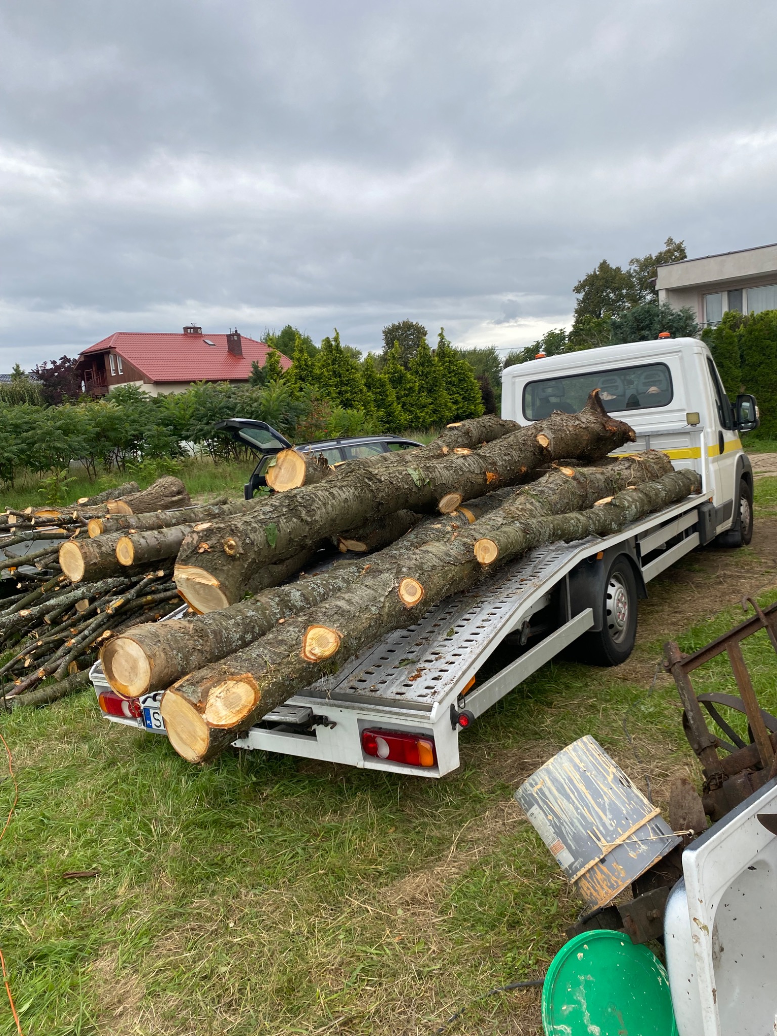 Przyczepa samochodu dostawczego załadowana pociętymi kłodami drewna i gałęziami, widoczne budynki mieszkalne w tle.