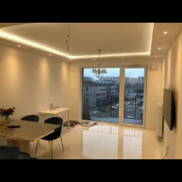 Nowoczesne wnętrze pokoju dziennego z białymi ścianami, podwieszanym sufitem z oświetleniem LED, dużym oknem balkonowym i minimalistycznym umeblowaniem, w trakcie wykańczania, widoczne kable od lampy.