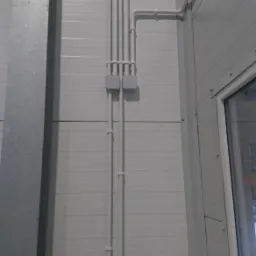 Białe rury instalacji elektrycznej prowadzone wzdłuż ściany z paneli w hali, widoczne puszki i gniazdo elektryczne przy podłodze.