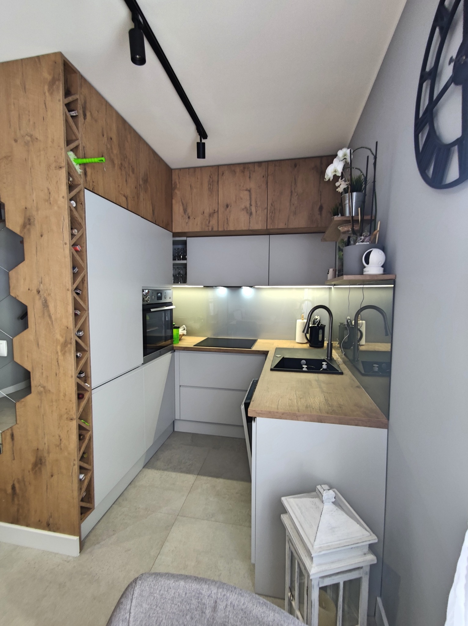 Nowoczesna, jasna kuchnia z szarymi szafkami, drewnianymi akcentami i czarnym oświetleniem szynowym. Regał na wino z boku, dekoracje na półkach i duży zegar na ścianie.