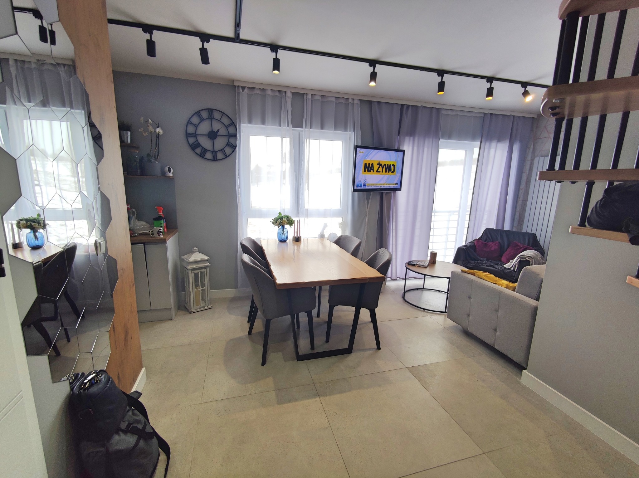 Jasne wnętrze salonu z drewnianym stołem i szarymi krzesłami, telewizor z napisem 'NA ŻYWO', podłoga wyłożona płytkami imitującymi kamień, oświetlenie punktowe na suficie.