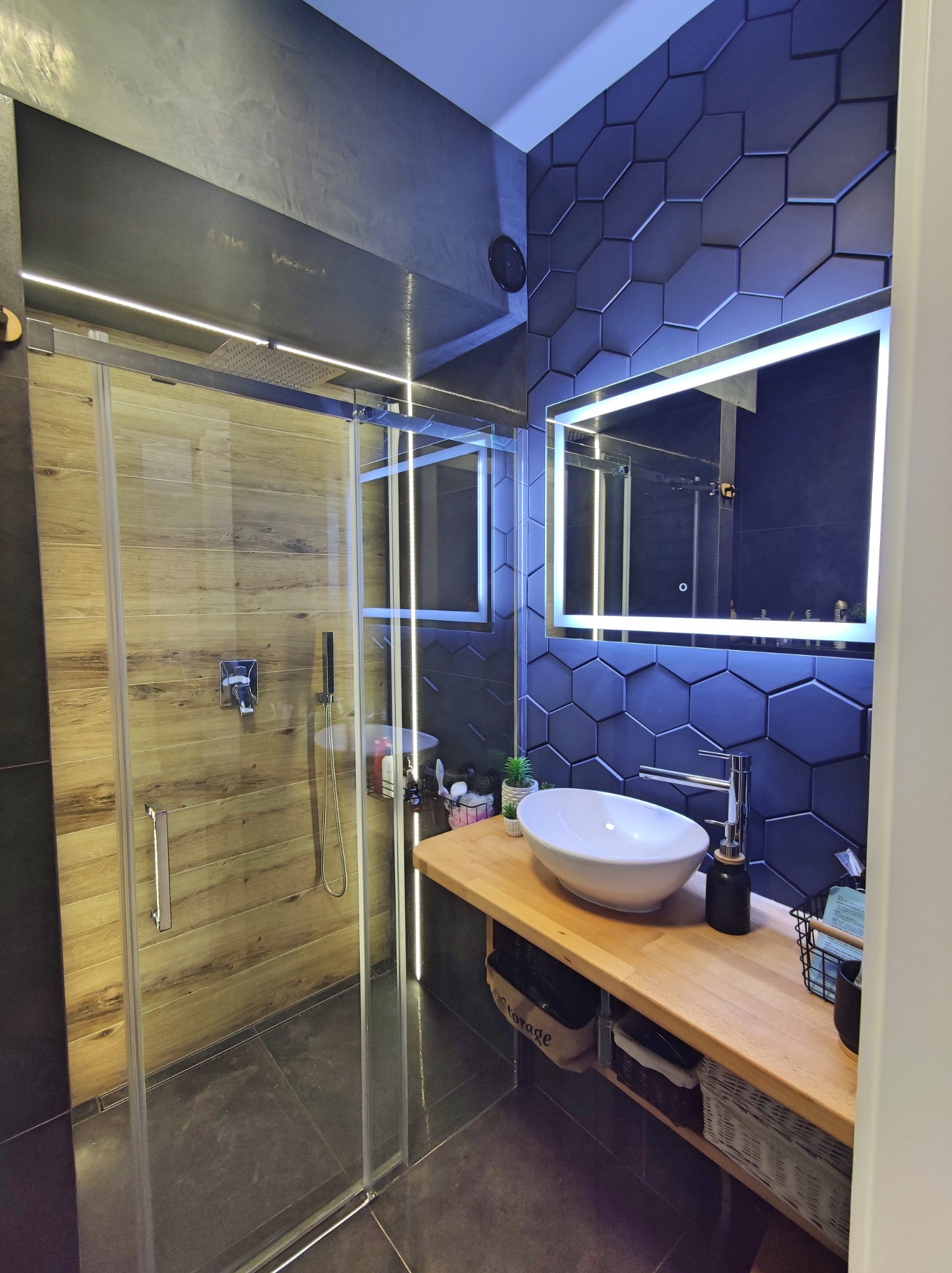 Nowoczesna łazienka z kabiną prysznicową typu walk-in, drewnopodobnymi płytkami wewnątrz, heksagonalnymi, ciemnymi płytkami na ścianie z podświetlanym lustrem, umywalką nablatową i drewnianym...