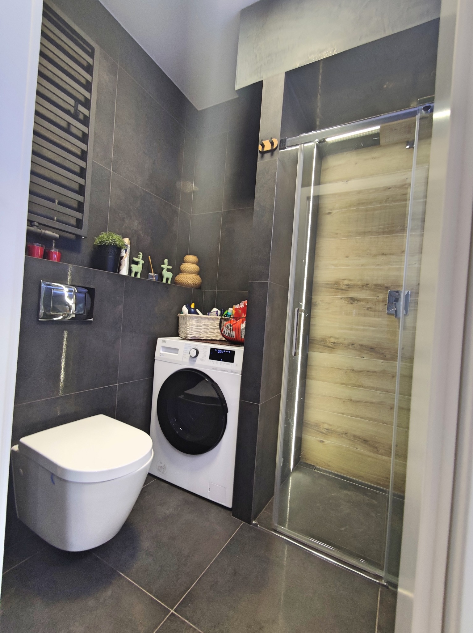 Nowoczesna łazienka z szarymi płytkami, podwieszaną toaletą, pralką i kabiną prysznicową z drewnopodobnymi panelami na ścianie, dekoracje na półce.