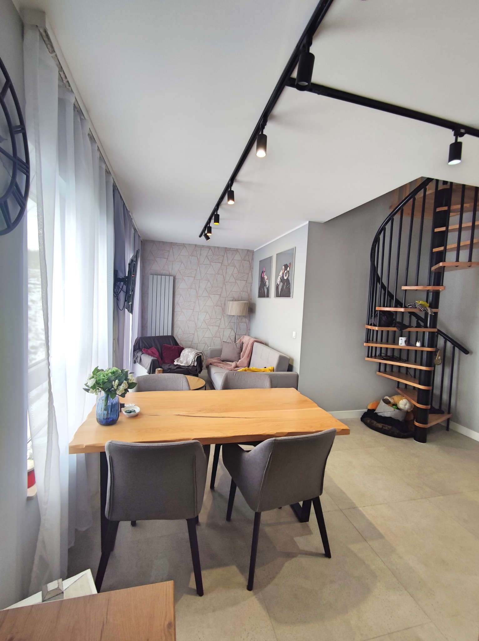 Jasne wnętrze pokoju dziennego z drewnianym stołem, szarymi krzesłami, spiralnymi schodami i geometryczną tapetą w tle.