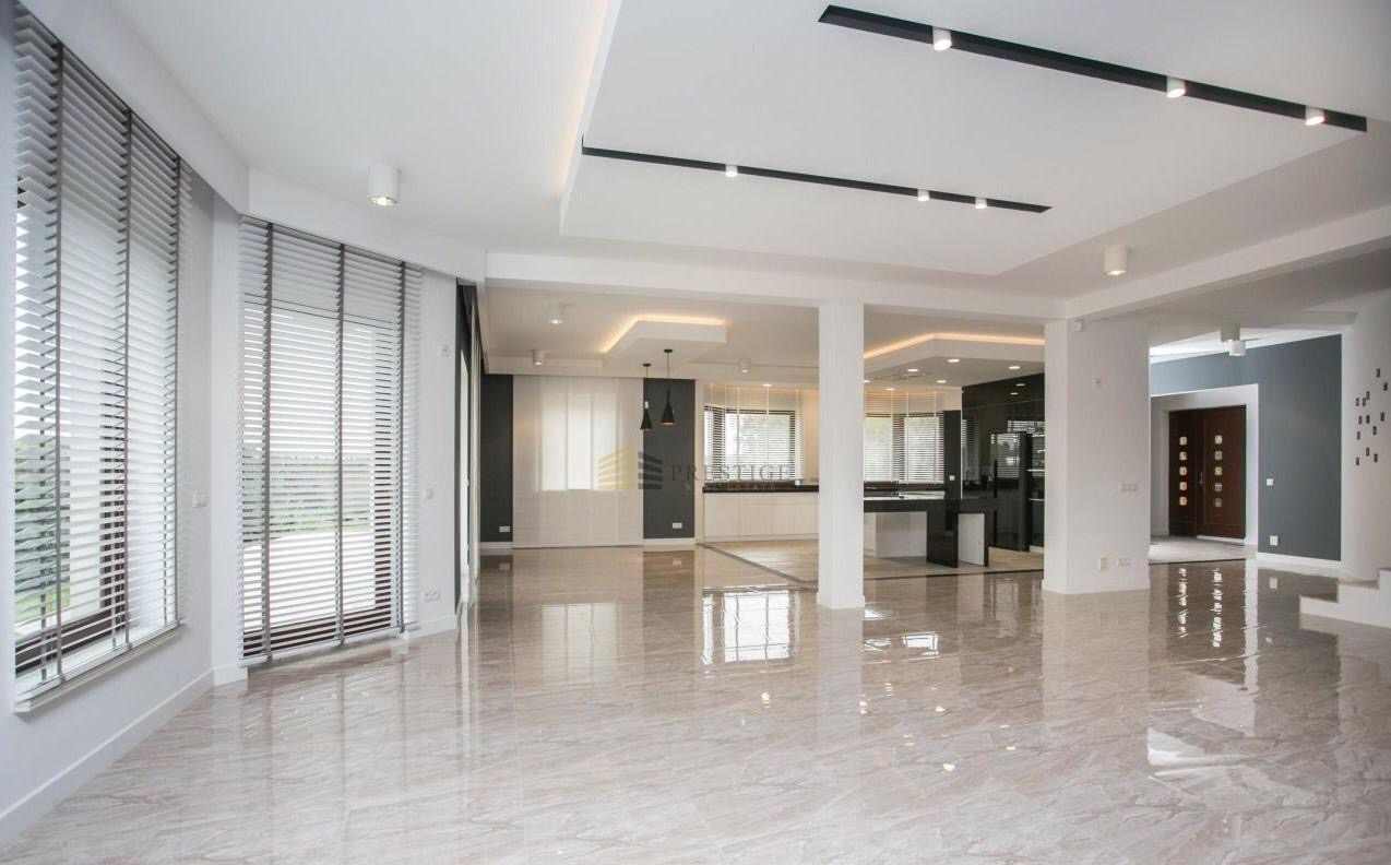 Przestronne wnętrze domu z marmurową podłogą, białymi ścianami, dużymi oknami z żaluzjami i nowoczesnym oświetleniem sufitowym.