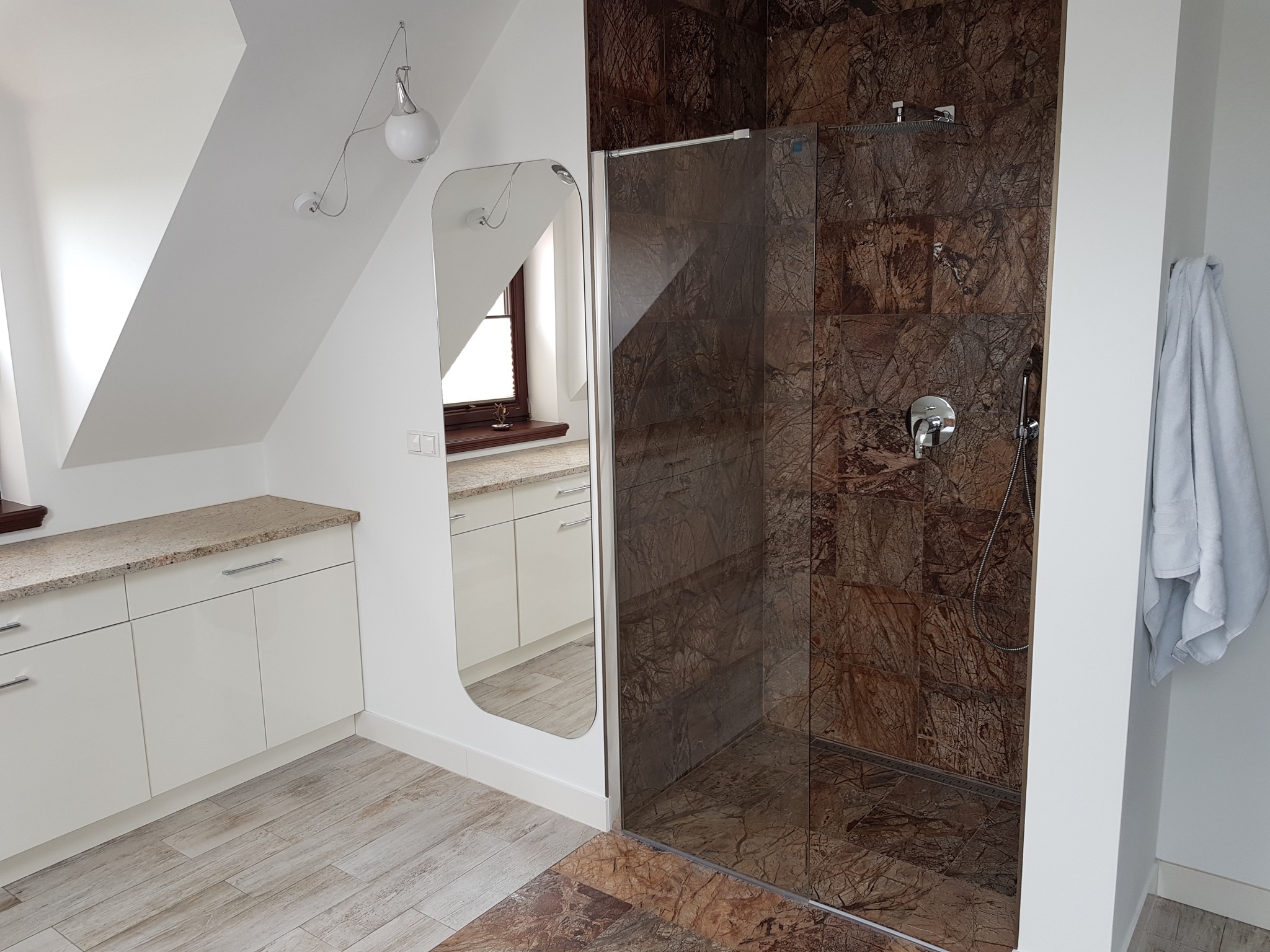 Nowoczesna łazienka na poddaszu z kabiną prysznicową wyłożoną brązowymi płytkami imitującymi marmur, białe meble łazienkowe z kamiennym blatem, duże lustro i drewnopodobna podłoga.