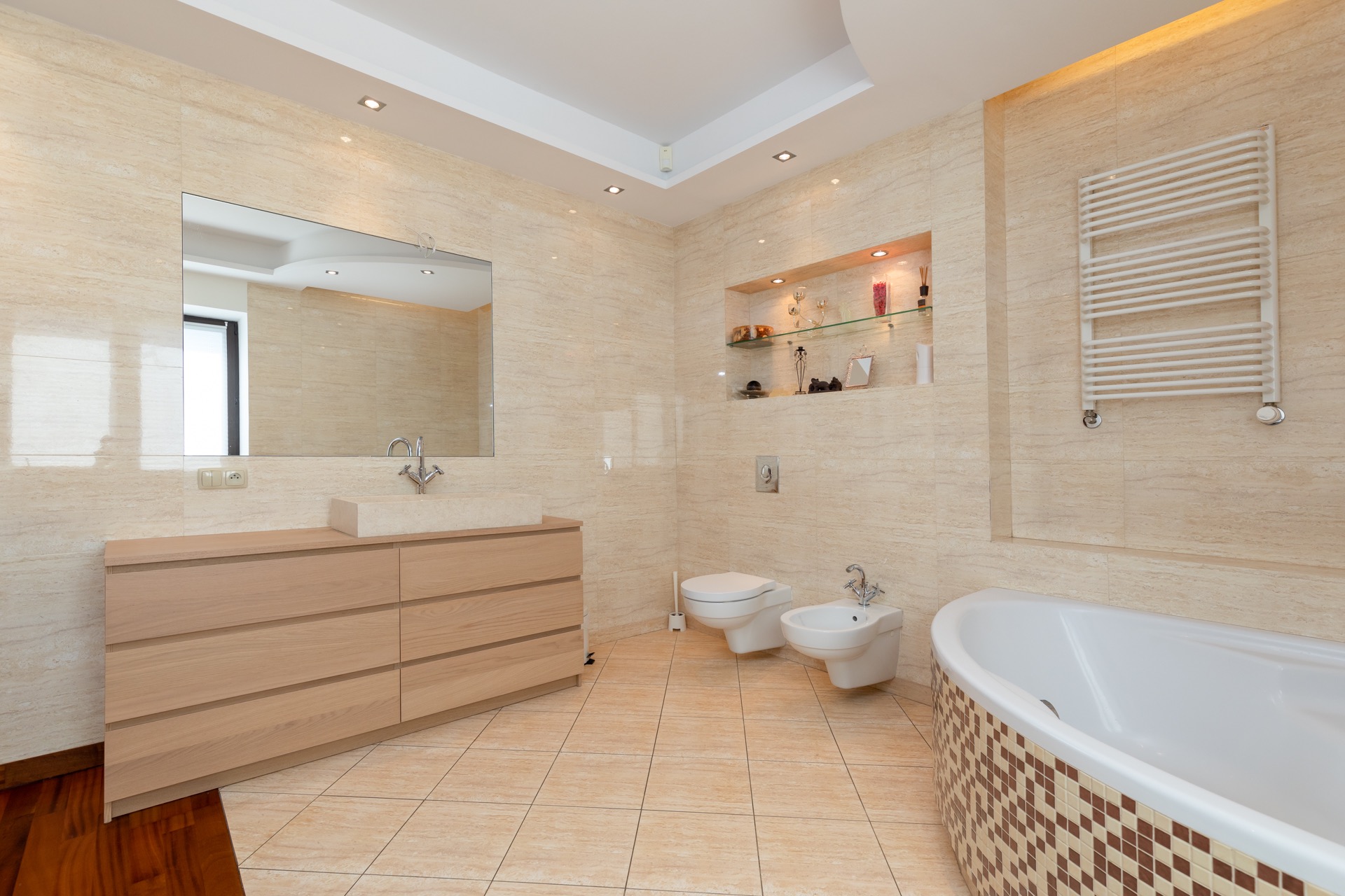 Jasna łazienka wykończona płytkami imitującymi trawertyn, z drewnianą szafką pod umywalką, wiszącą toaletą i bidetem oraz wanną z mozaikową obudową.