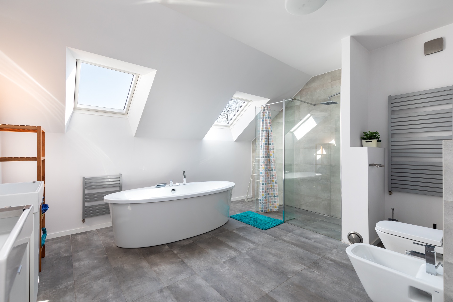 Jasna łazienka na poddaszu z oknami dachowymi, owalną wanną, kabiną prysznicową ze wzorzystą zasłoną, szarą podłogą imitującą kamień i białymi sanitariatami.