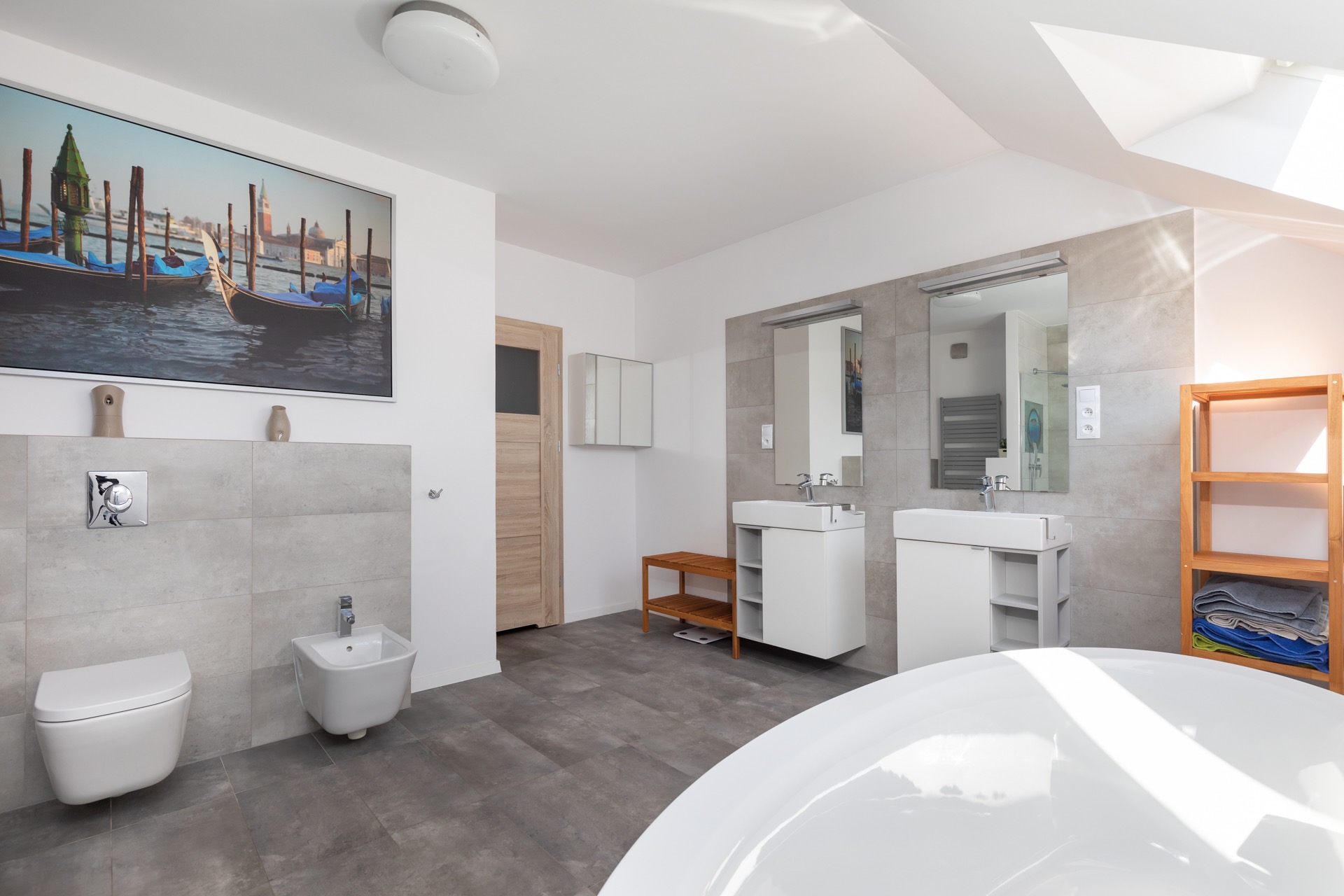 Jasna łazienka z szarymi płytkami, dwiema umywalkami, toaletą, bidetem, drewnianym regałem i obrazem z motywem weneckim.