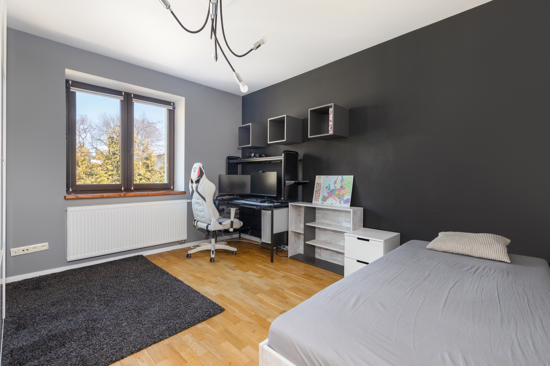 Pokój młodzieżowy z szarą ścianą, biurkiem gamingowym, białym krzesłem, drewnianą podłogą i łóżkiem z szarą pościelą, widziany w jasnym świetle dziennym.