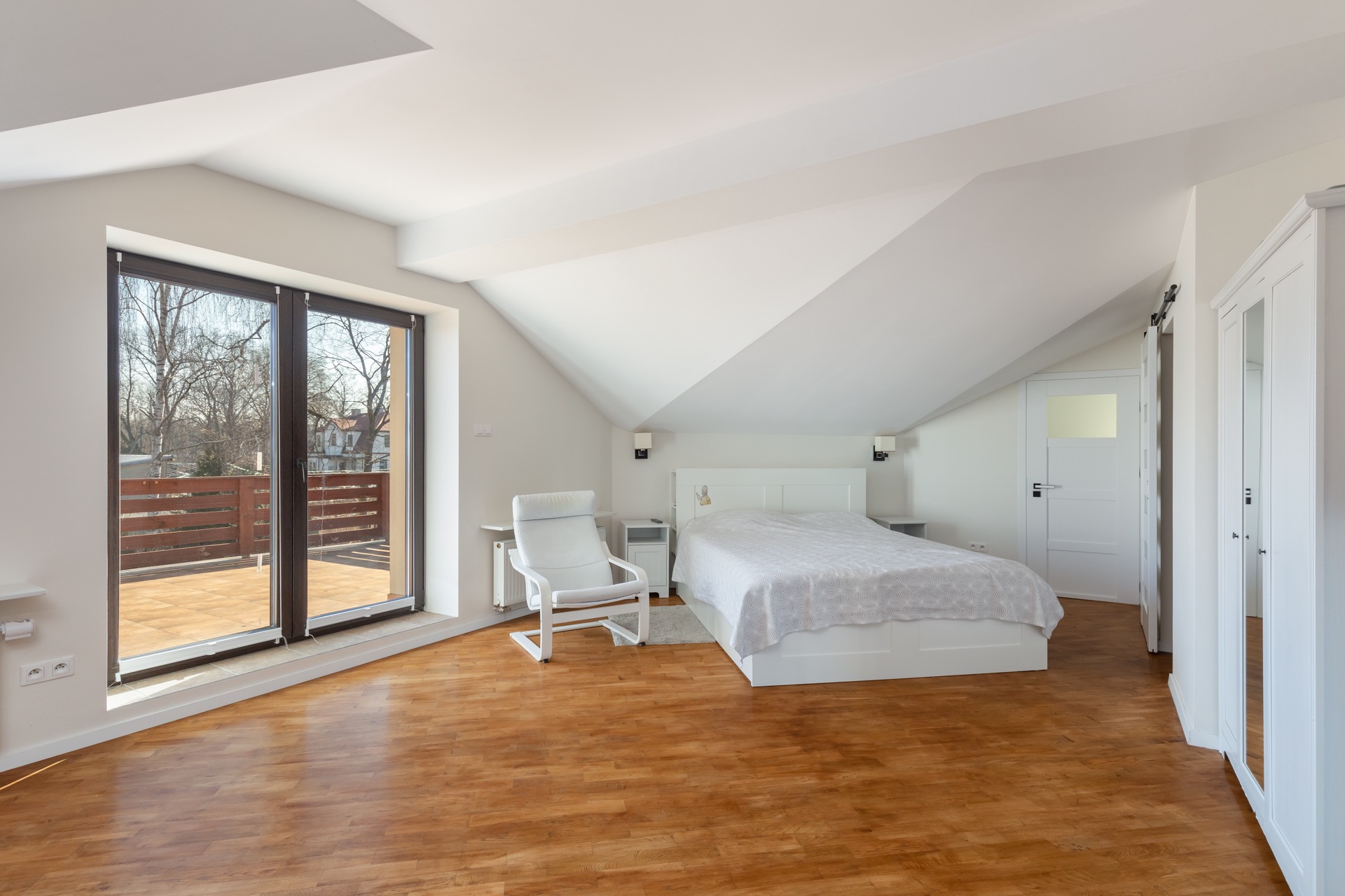 Jasne, przestronne poddasze z drewnianą podłogą, białymi ścianami, oknem balkonowym i minimalistycznym umeblowaniem, w tym białe łóżko, szafa i fotel.