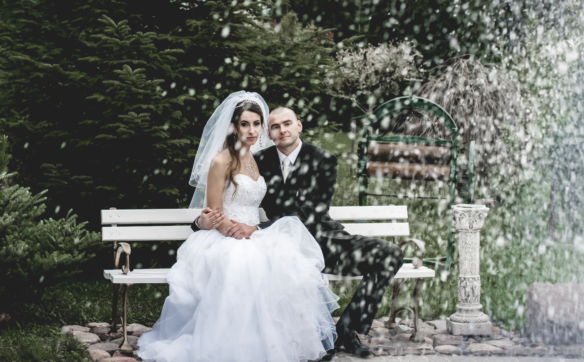 Para młoda siedzi na białej ławce na tle zieleni i fontanny, panna młoda w białej sukni z tiarą i welonem, pan młody w czarnym garniturze, oboje patrzą w kamerę.