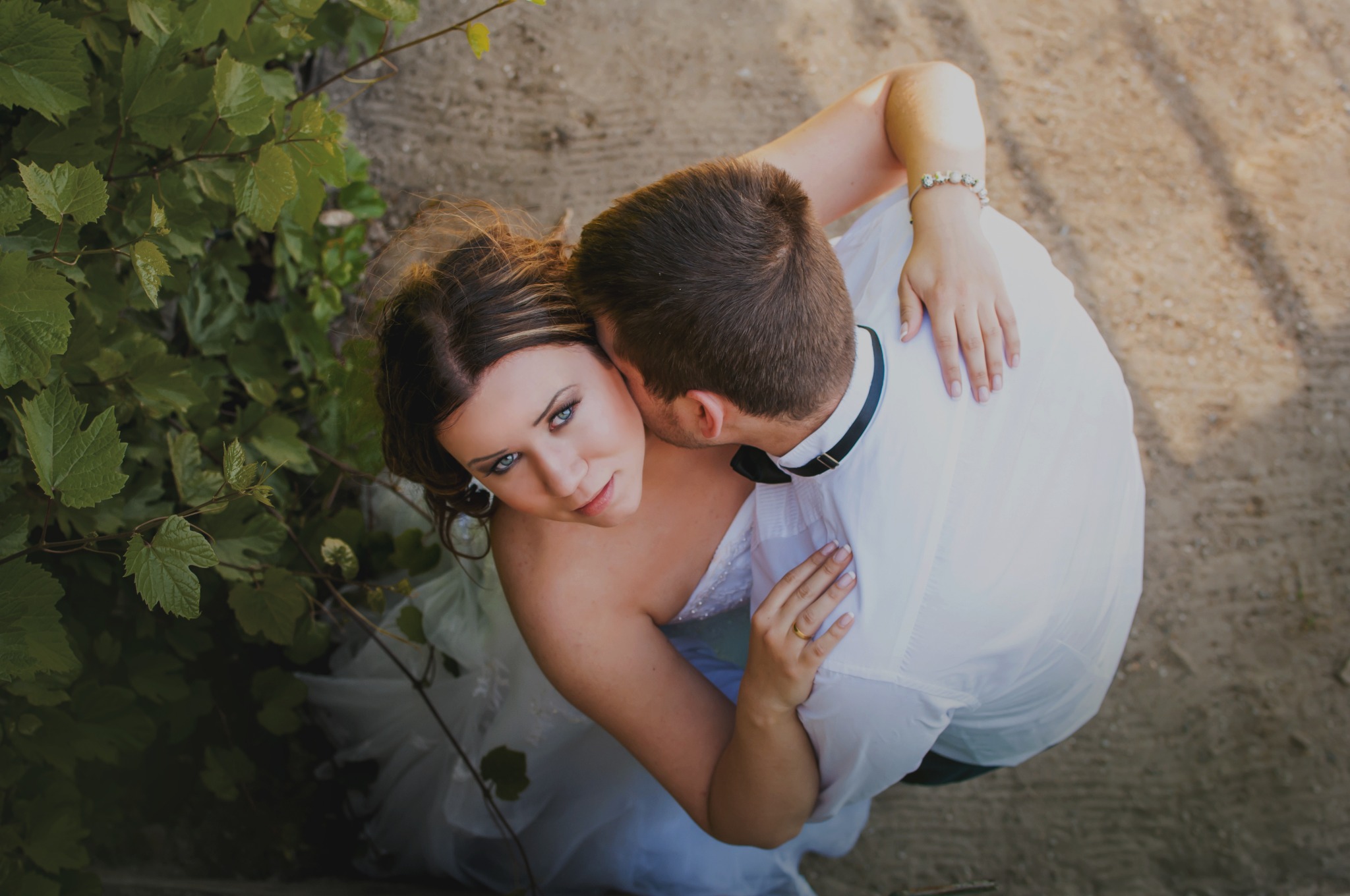 Para młoda w objęciach, widok z góry, panna młoda z wyrazistym makijażem oczu spogląda w obiektyw, a pan młody w białej koszuli z muchą przytula ją, tło stanowią zielone liście i piaszczysta ziemia.