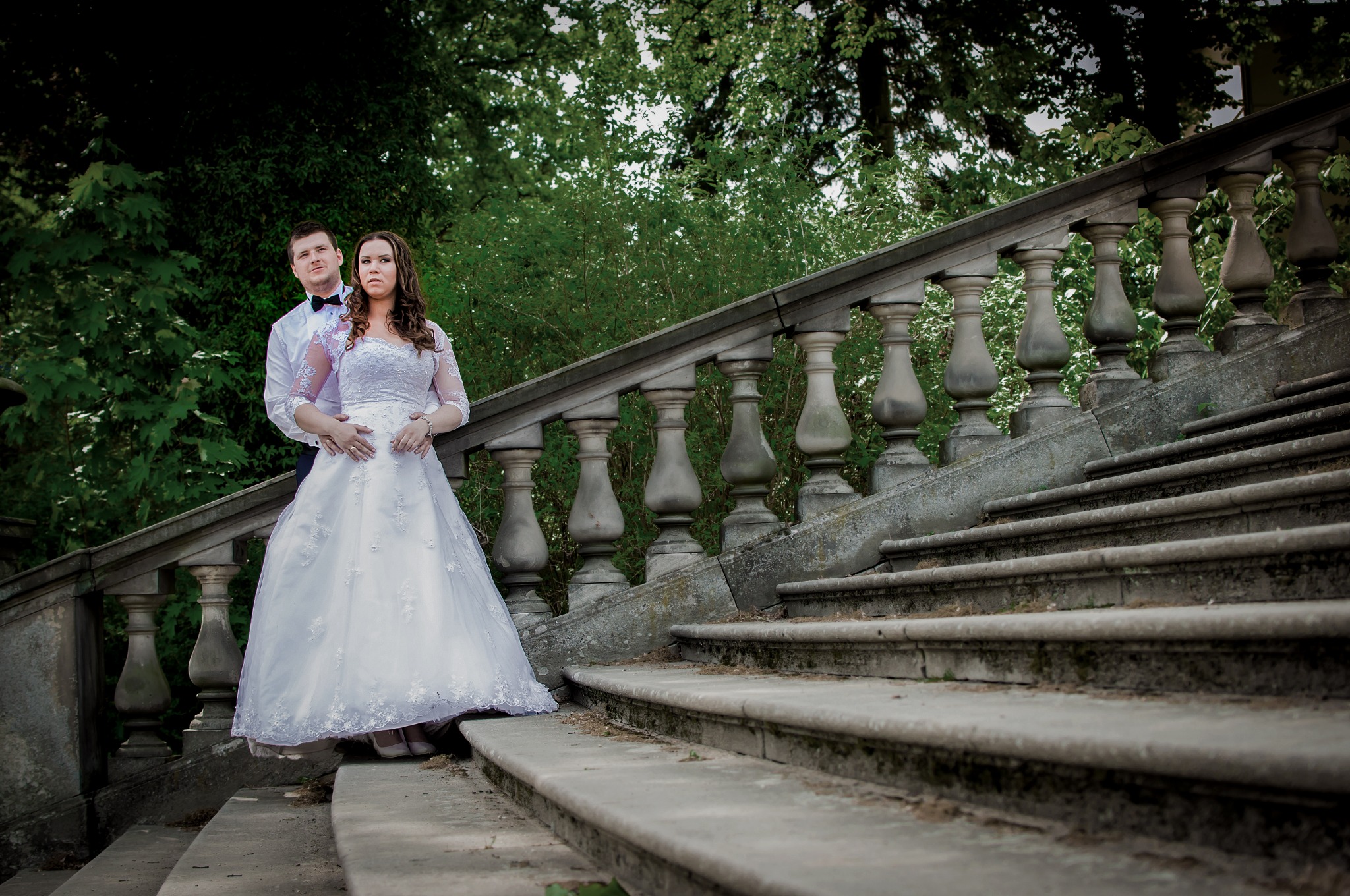 Para w strojach ślubnych pozuje na starych kamiennych schodach z balustradą, otoczonych zielenią.