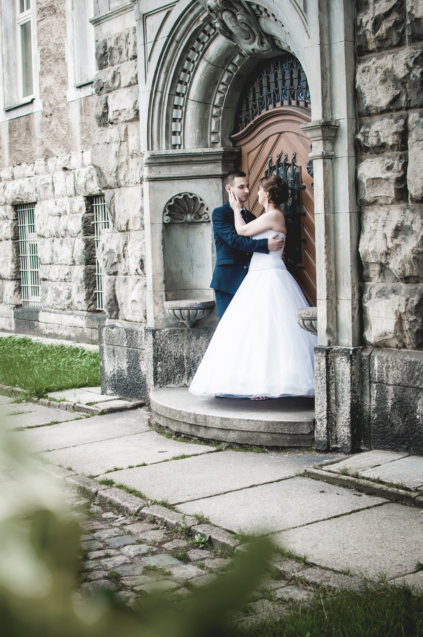 Para młoda w objęciach na tle kamiennej fasady budynku z łukowym wejściem i drewnianymi drzwiami z metalowymi zdobieniami.