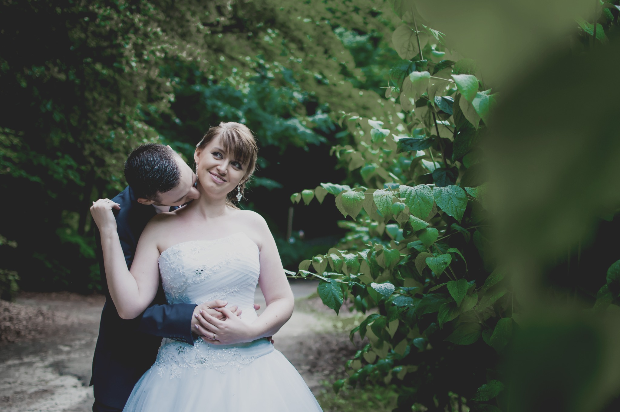 Panna młoda w białej sukni, obejmowana przez pana młodego, który delikatnie całuje ją w szyję na tle zieleni.