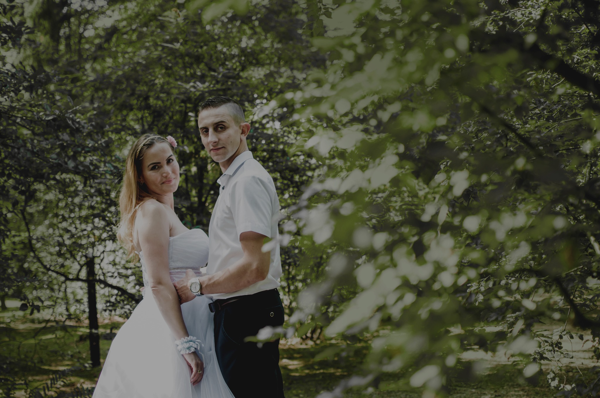 Para w strojach ślubnych pozuje na tle zieleni, mężczyzna obejmuje kobietę w talii, oboje patrzą w obiektyw.