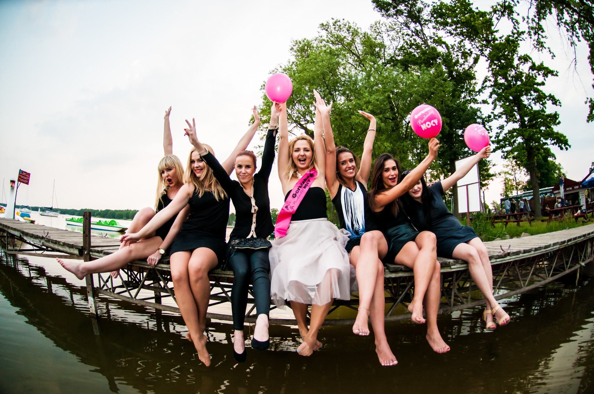 Grupa sześciu uśmiechniętych kobiet siedzi na drewnianym pomoście nad wodą, trzymając różowe balony z napisem; jedna z kobiet ma różową szarfę z napisem 'Gorączka Panieńska Nocy'.