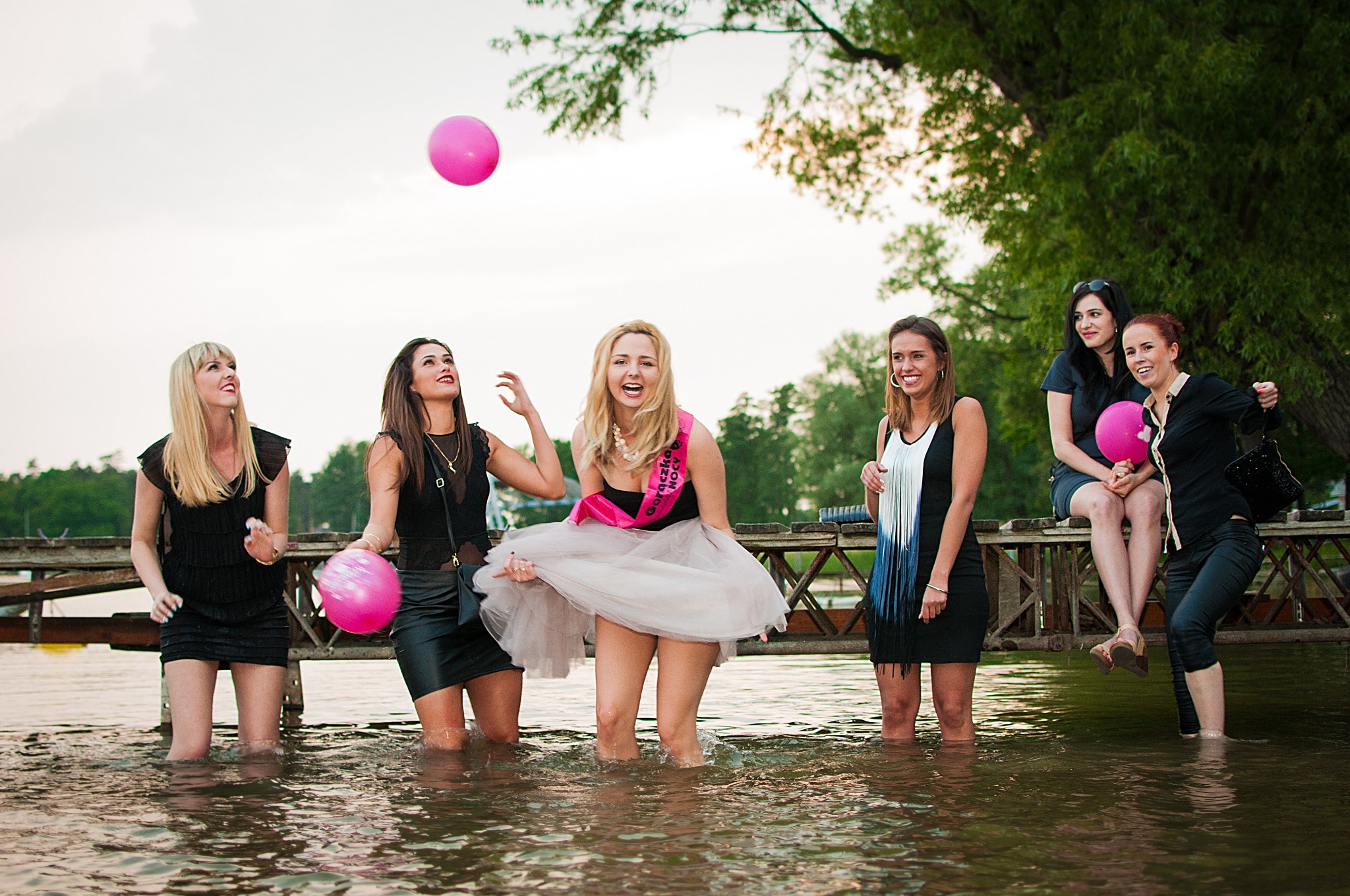 Szczęśliwe kobiety w czarnych sukienkach bawią się w wodzie podczas wieczoru panieńskiego w Szczecinie, jedna z nich ma różową szarfę z napisem, a w tle widać drewniany pomost i drzewa.