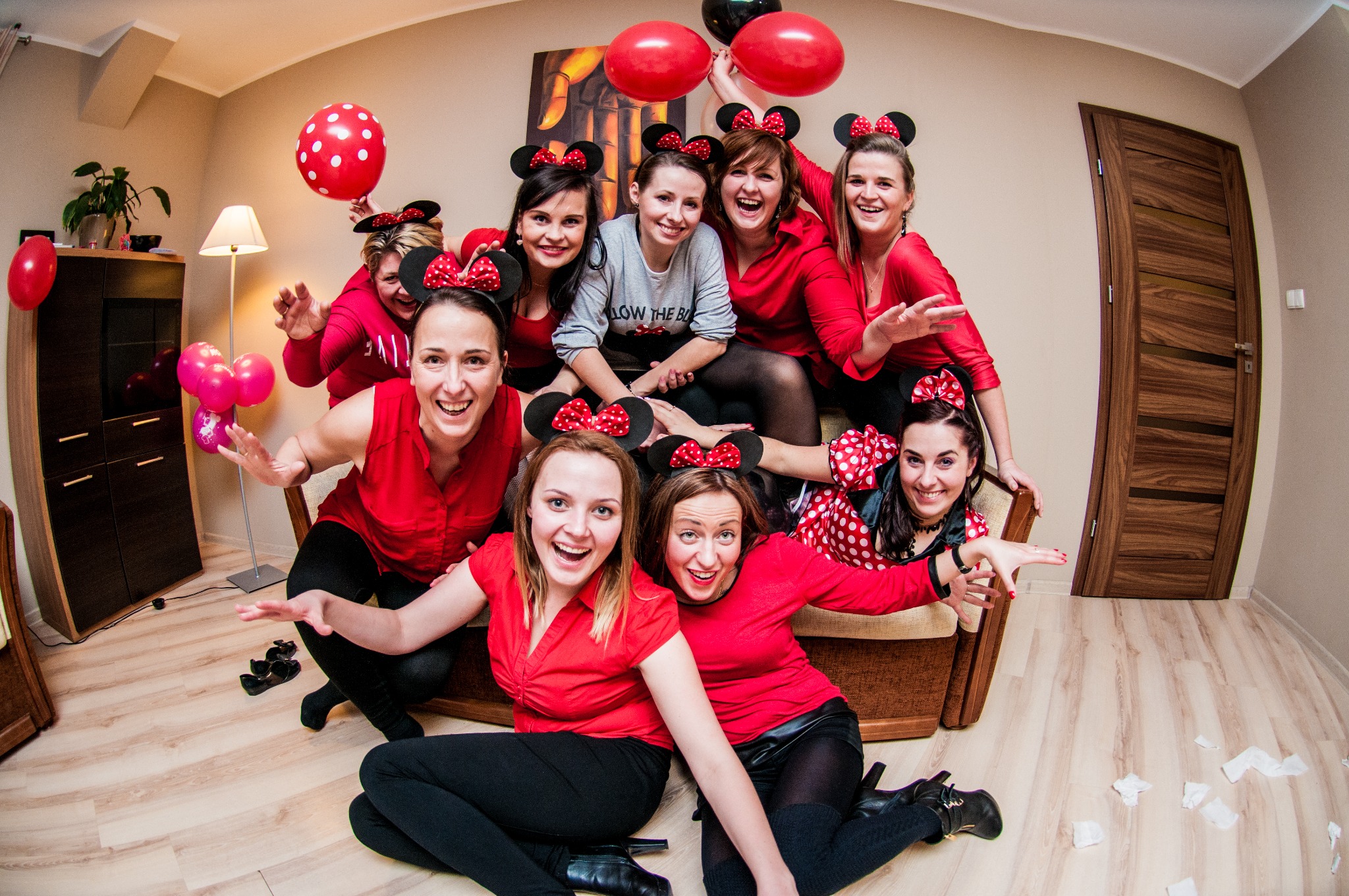 Grupa uśmiechniętych kobiet w czerwonych bluzkach i opaskach z uszami Myszki Miki pozuje w pomieszczeniu z balonami, radosna atmosfera.