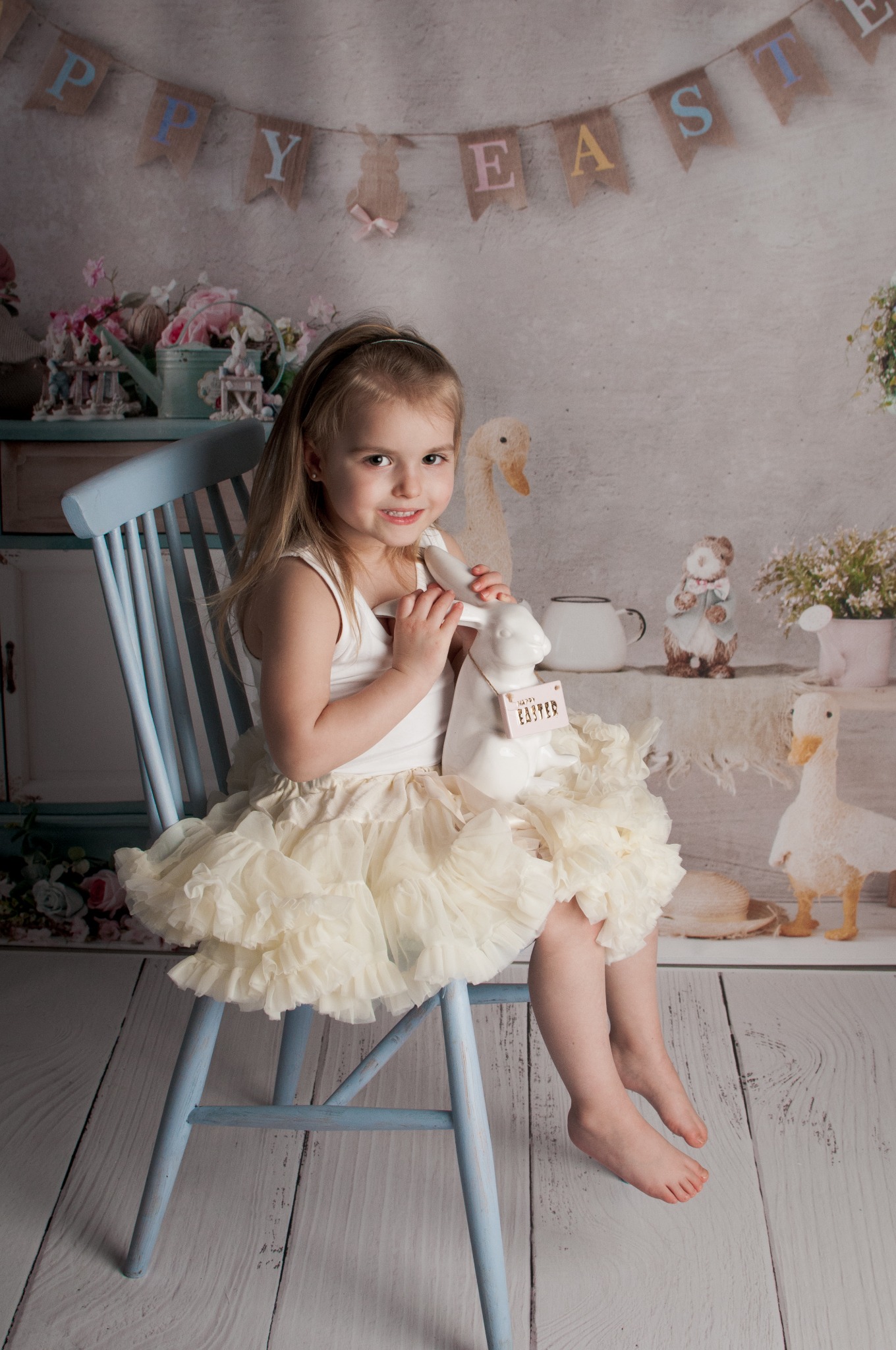 Dziewczynka w kremowej spódniczce siedzi na błękitnym krześle, trzymając białego ceramicznego zająca z napisem 'Happy Easter', na tle dekoracji wielkanocnych.