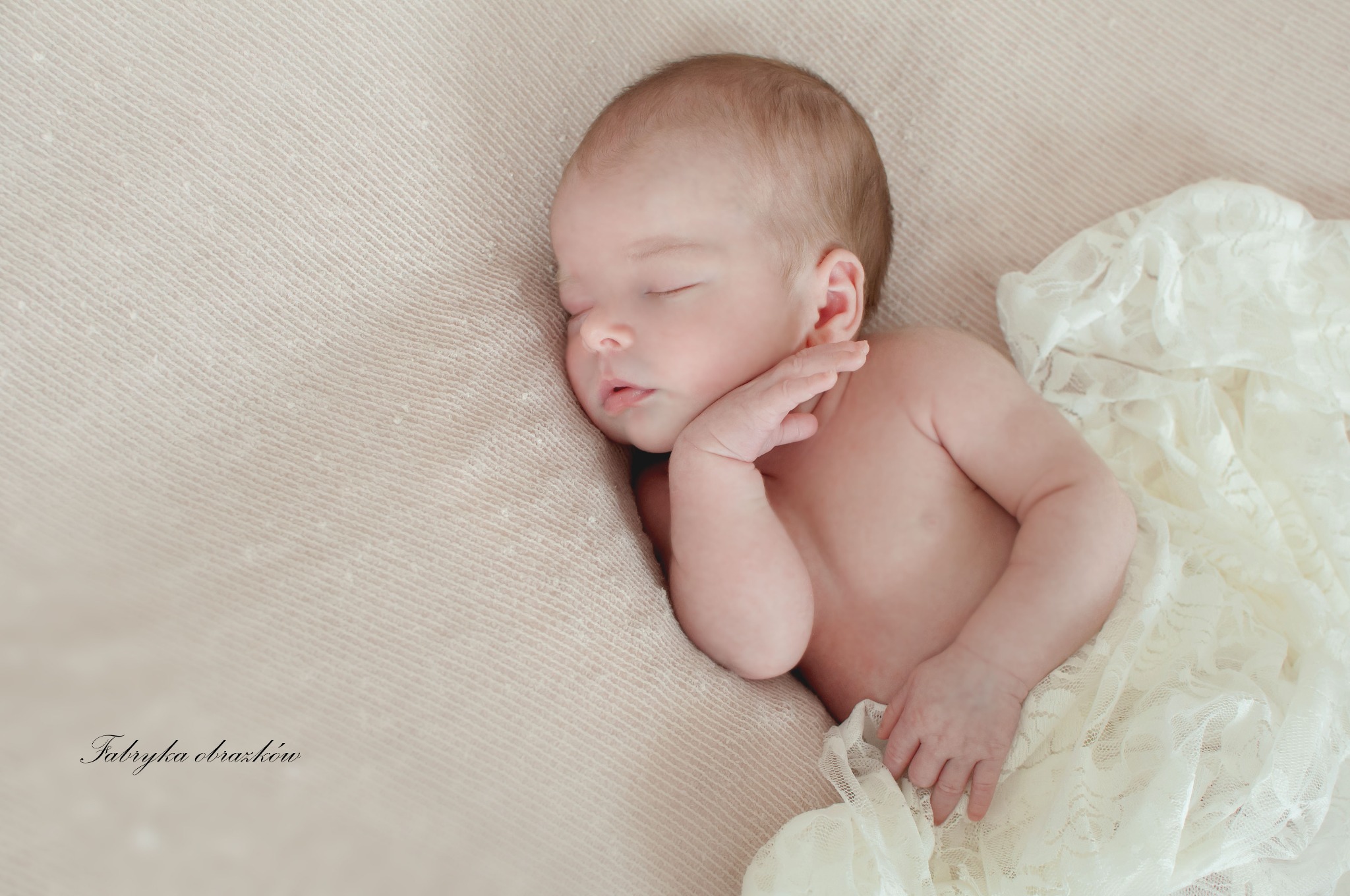 Śpiący noworodek, ułożony na beżowym tle z koronkową tkaniną, podpierający główkę małą rączką.