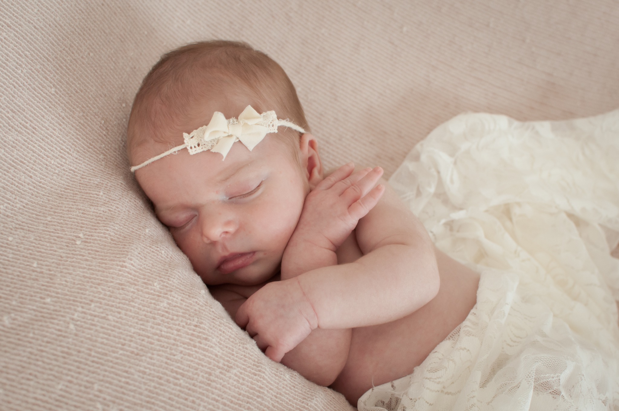 Śpiąca noworodka z kremową opaską z kokardką, leżąca na beżowym tle z koronkową tkaniną.