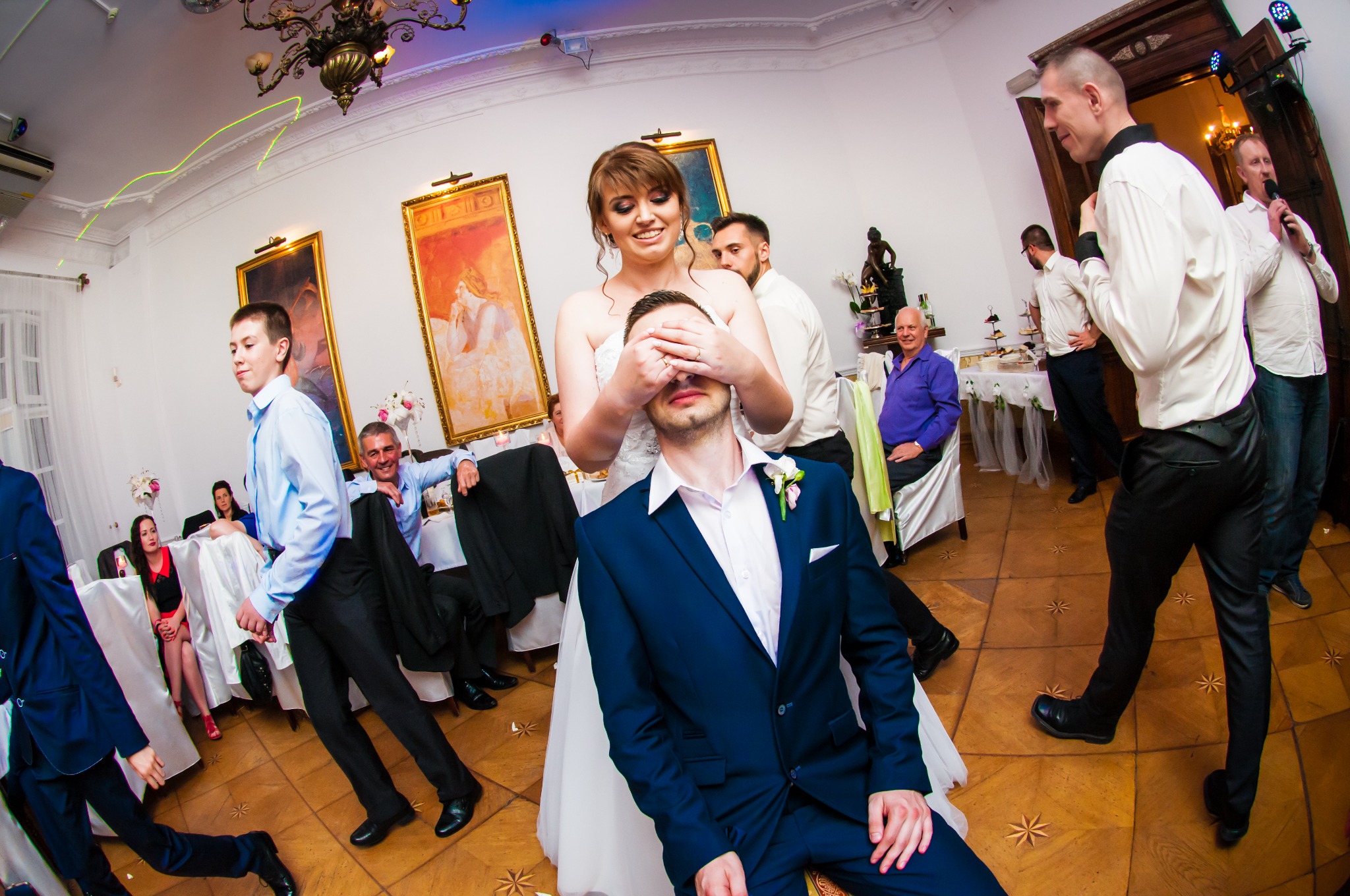 Panna młoda zakrywa oczy siedzącemu panu młodemu podczas wesela, w tle goście i sala z dekoracjami.