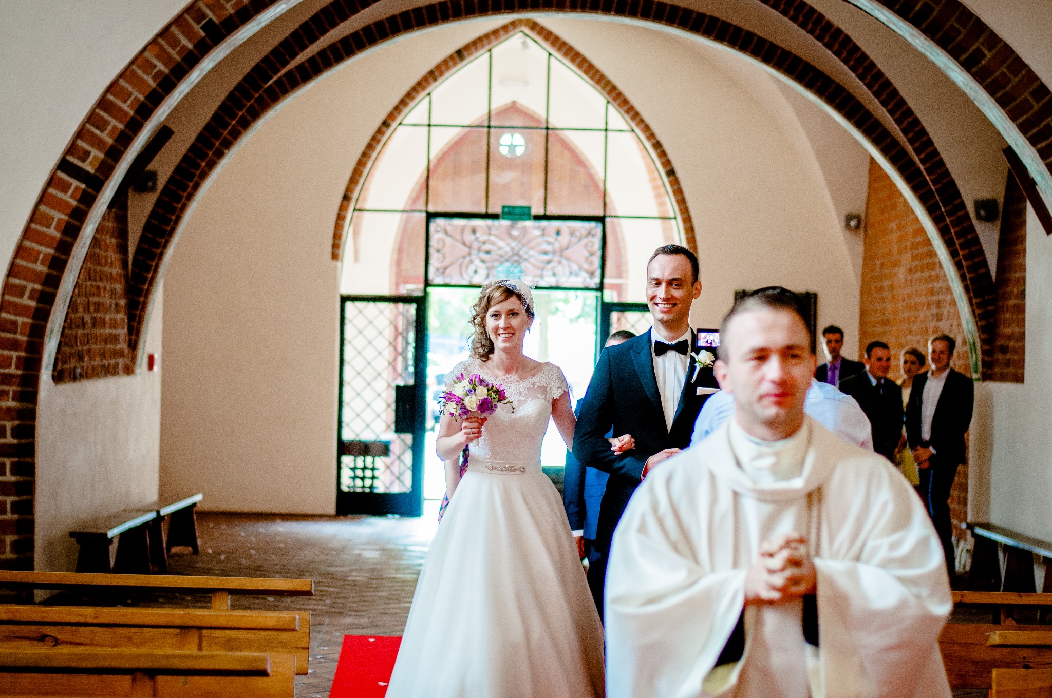 Para młoda wychodzi z kościoła, trzymając się za ręce, w tle ceglane łuki i goście.