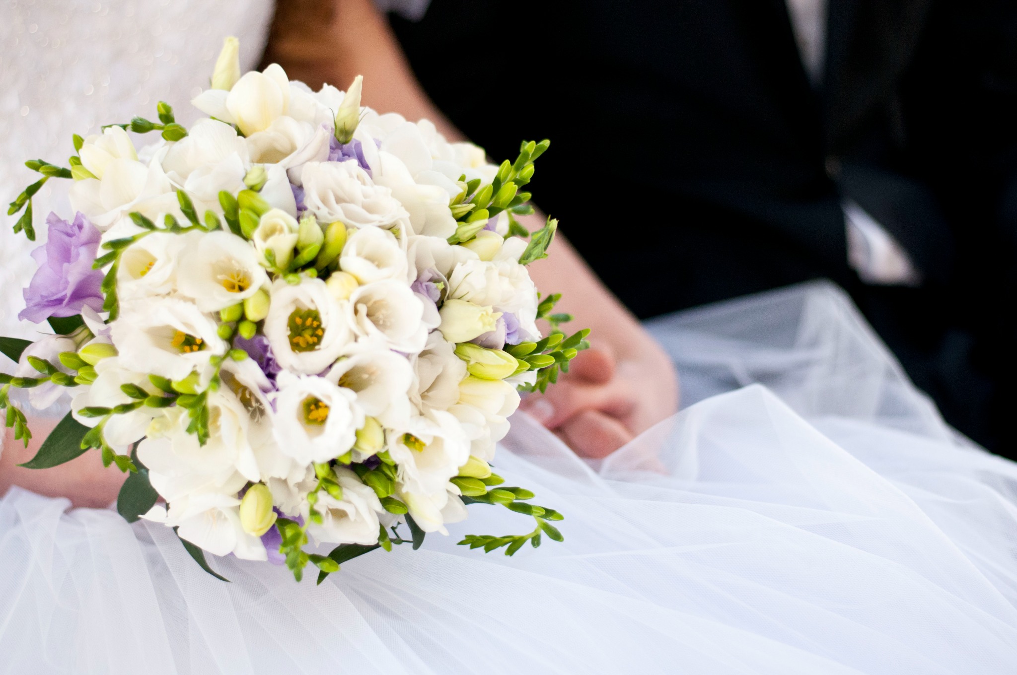 Panna młoda trzyma bukiet białych i fioletowych kwiatów na tle białej sukni i dłoni pana młodego.