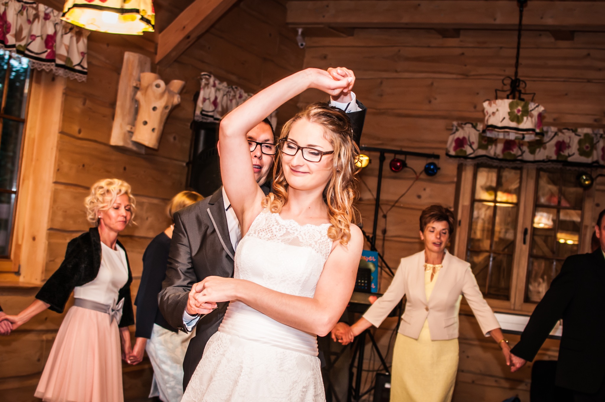 Para młoda tańczy na weselu w drewnianej sali, goście trzymają się za ręce w tle, dekoracje kwiatowe na lampach.