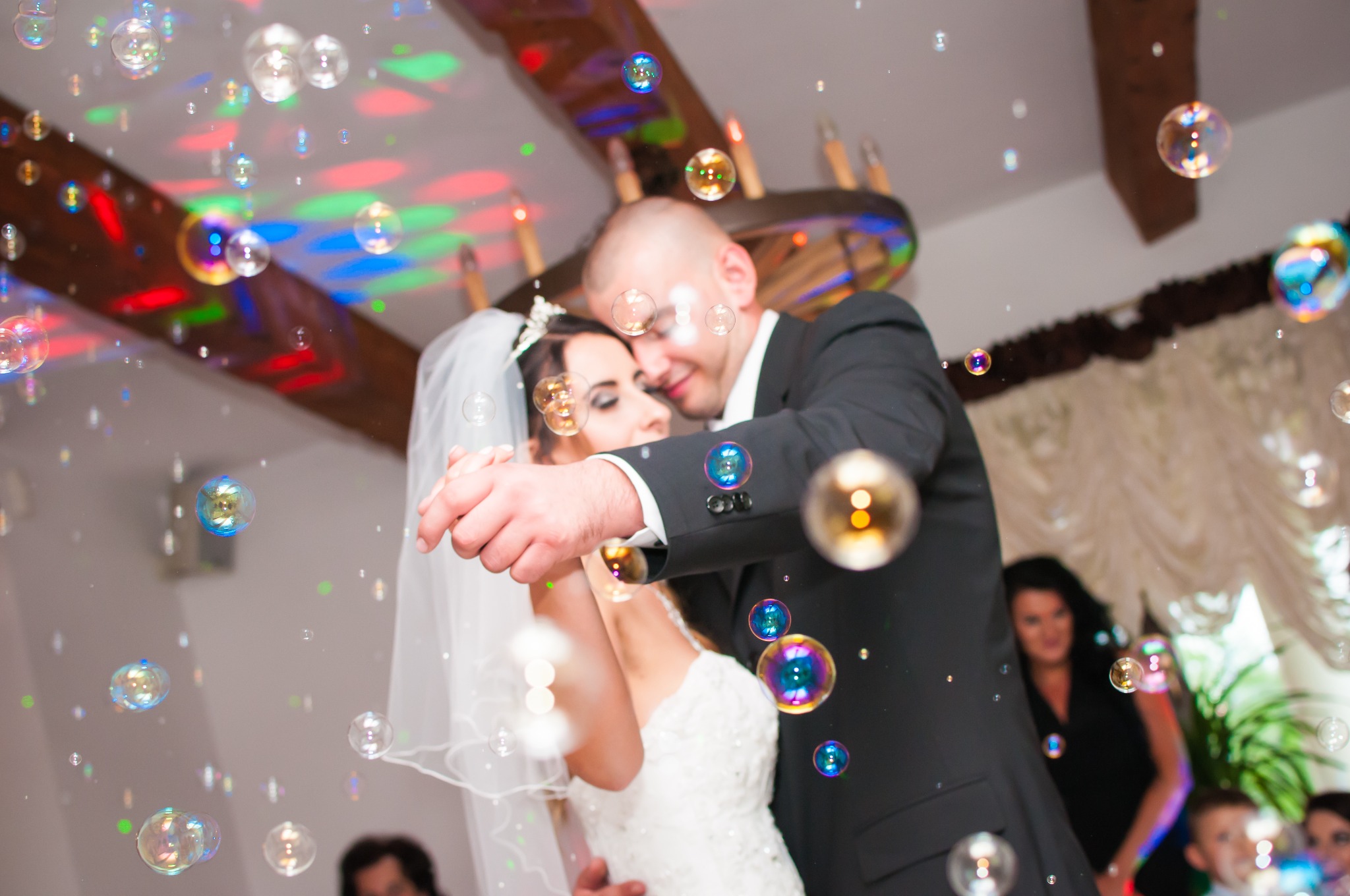 Para młoda w tańcu otoczona bańkami mydlanymi, ona w białej sukni i welonie z tiarą, on w czarnym garniturze, oświetlenie dyskotekowe w tle.