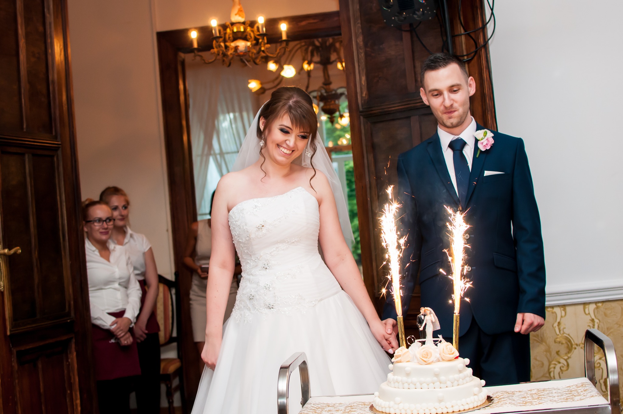 Uśmiechnięta panna młoda w białej sukni i welonie, wraz z panem młodym w granatowym garniturze, kroją tort weselny z fontannami iskier, w tle widoczne osoby obsługi.