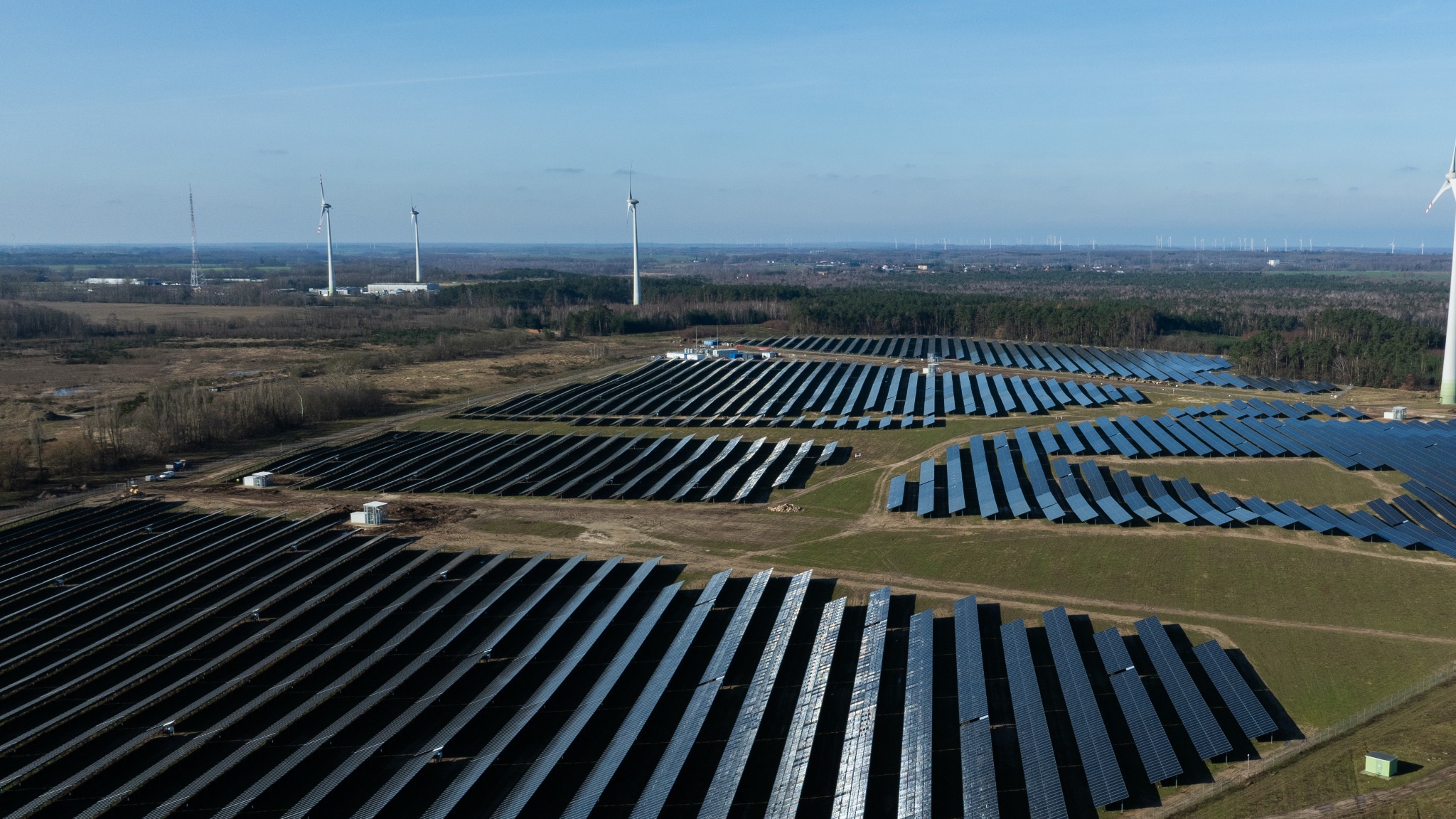 Rozległa farma fotowoltaiczna z rzędami paneli słonecznych, widok z góry, w tle turbiny wiatrowe i las.