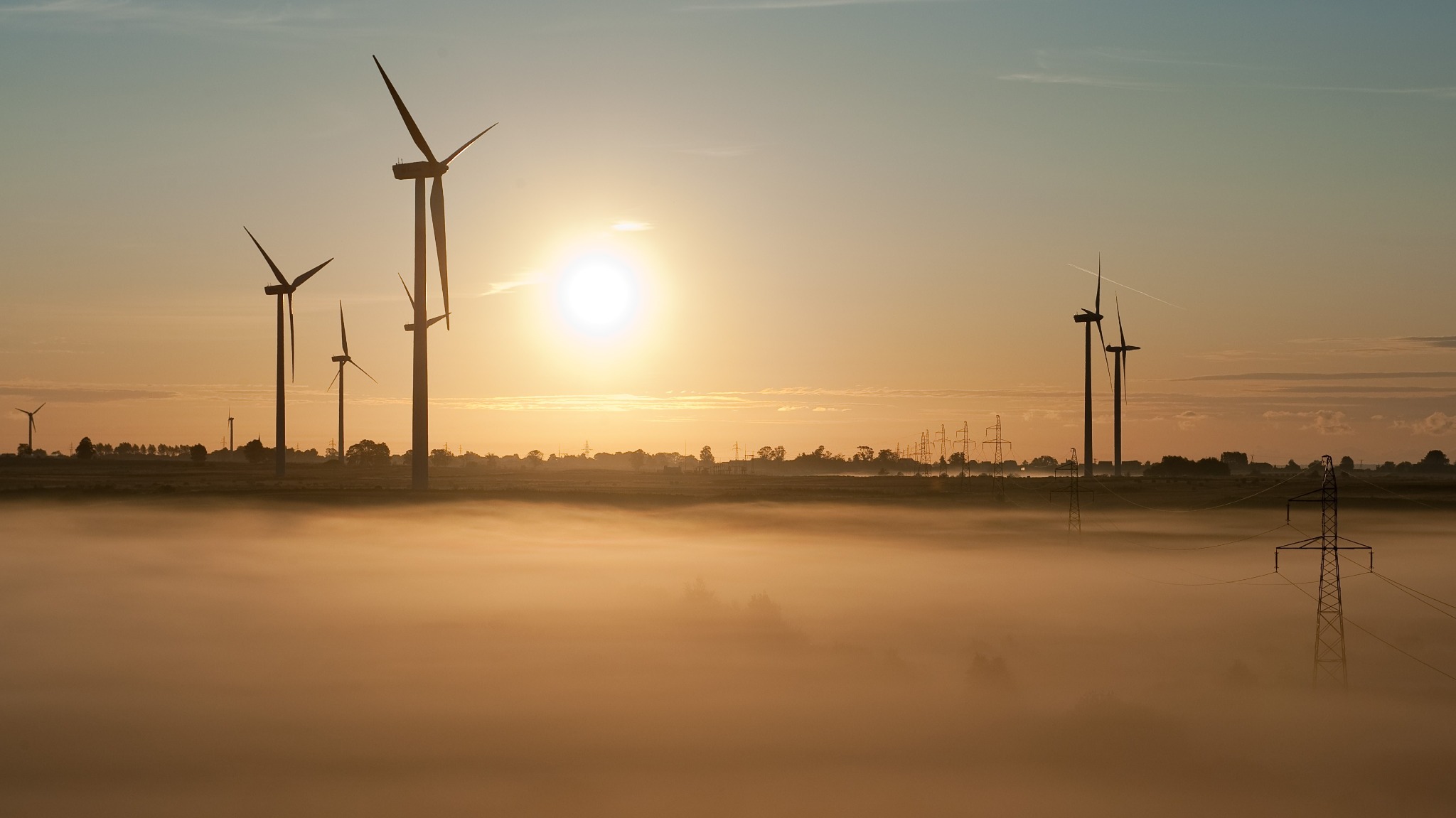 Poranna mgła otula farmę wiatrową z turbinami Gamesa G87, gdzie w tle widoczne są słupy wysokiego napięcia, a słońce wschodzi, tworząc złocistą poświatę.