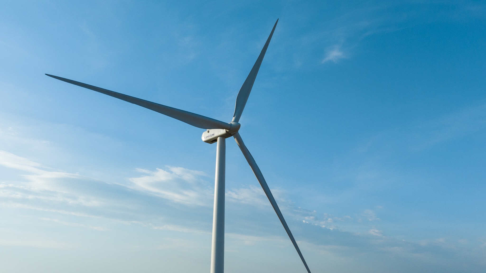 Farmy wiatrowe: Turbina wiatrowa Vestas z trzema łopatami na tle błękitnego nieba z delikatnymi obłokami, widoczna z dołu.